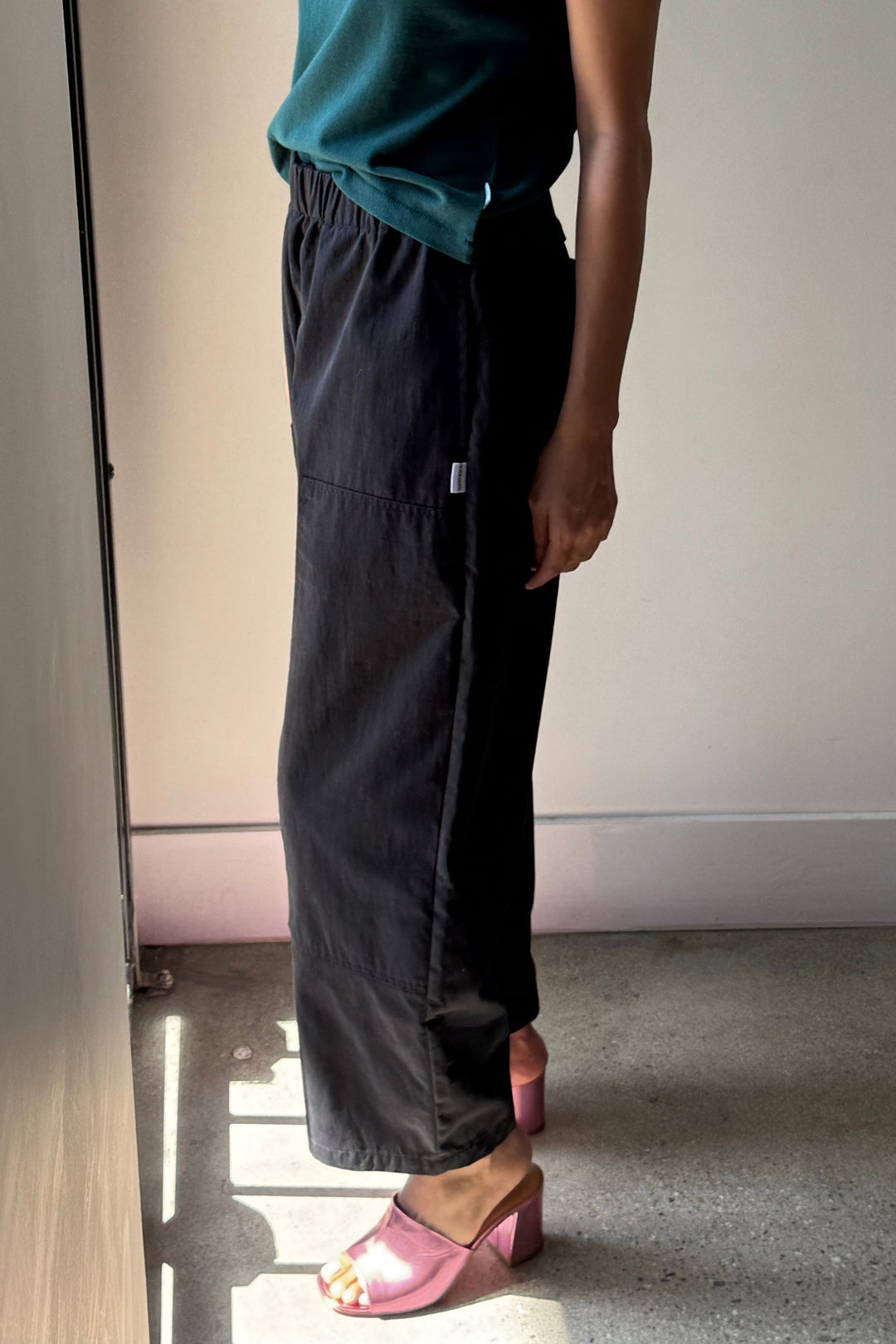 A person stands indoors in sunlight, wearing a green top and Le Bon Shoppe's Gardener's Pant in Black—crafted from lightweight cotton-nylon for a relaxed silhouette. Shiny pink open-toe heeled sandals complete the look, with only their torso and legs visible.
