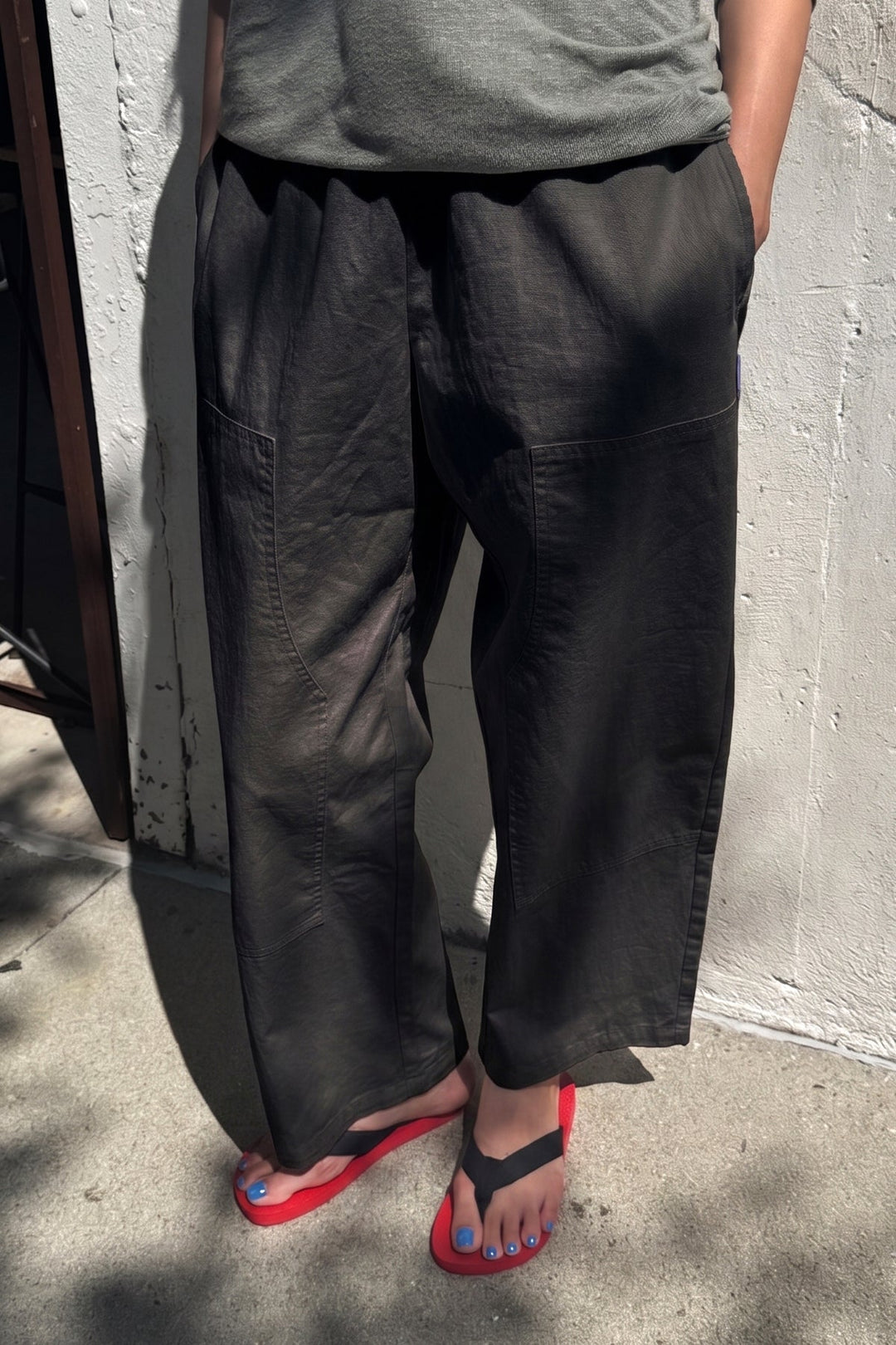 A person stands on the sidewalk with hands in their pockets, wearing Le Bon Shoppe - Gardener's Pant in Black from Le Bon Shoppe, which features a relaxed silhouette. They pair it with a gray shirt, black flip-flops with bright red soles, and blue nail polish. Sunlight casts shadows on the ground around them.