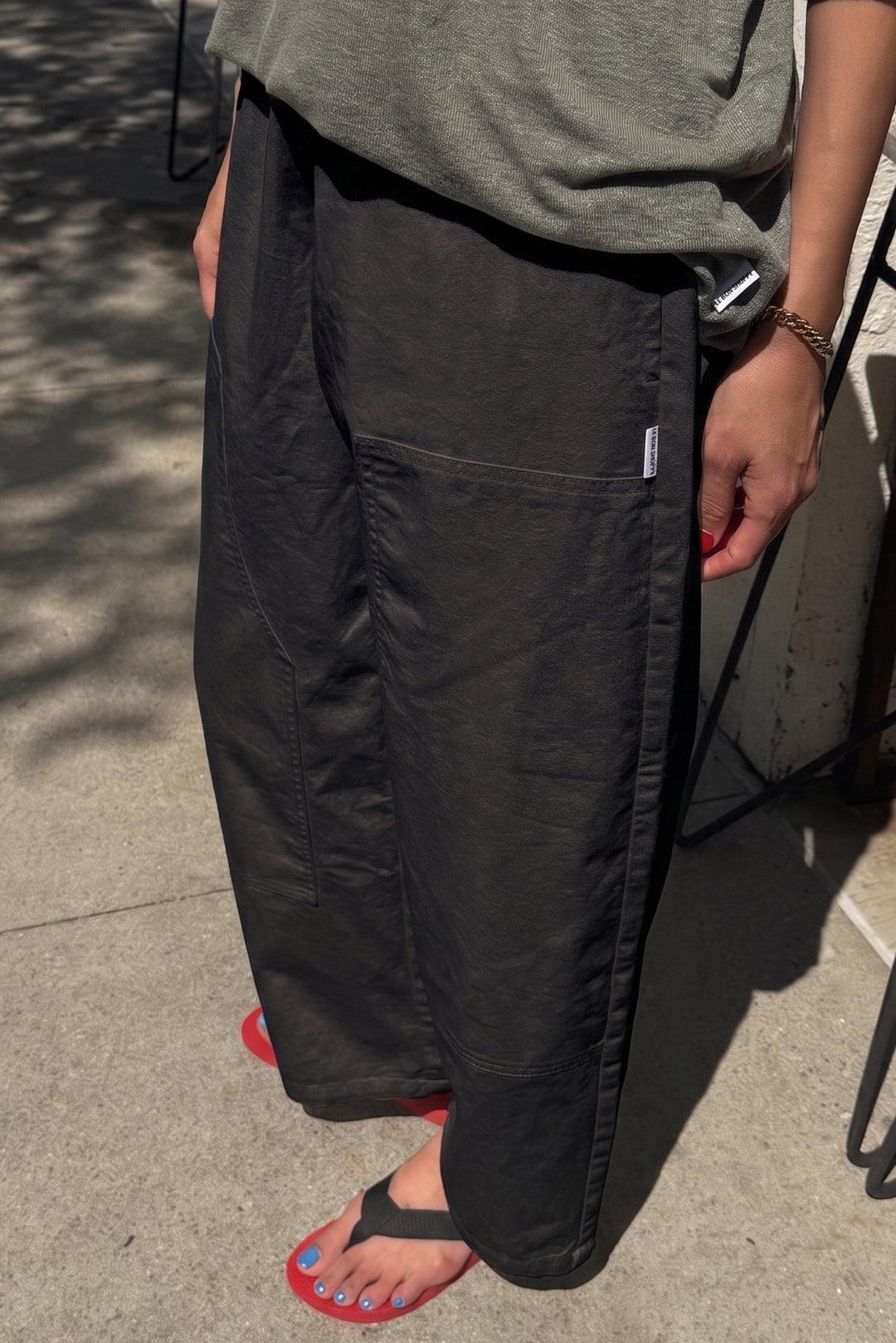 A person stands on a sunlit sidewalk wearing Le Bon Shoppe's Gardener's Pant in Black, crafted from lightweight cotton-nylon for a relaxed silhouette. Paired with a green top and black flip-flops, their blue-painted toenails and red-painted fingernails add pops of color. Only the lower half of the body is visible, drawing attention to the easy fit of Le Bon Shoppe’s signature pants.