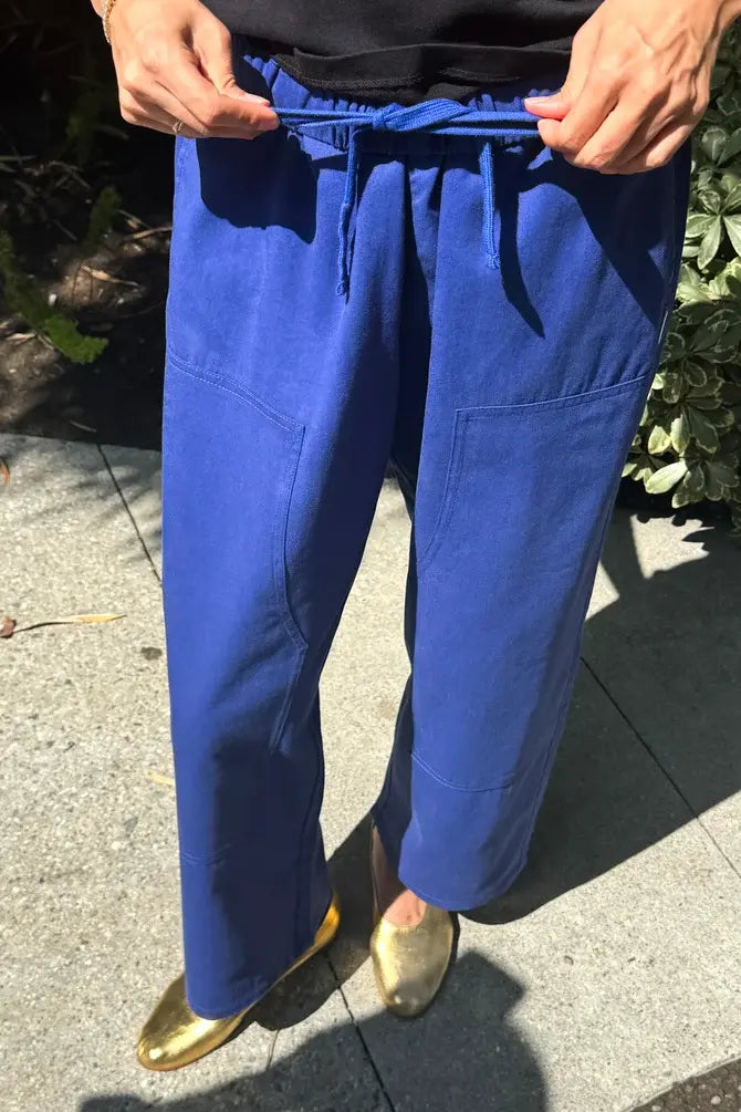 A person is wearing Le Bon Shoppe's Gardener's Pant in Deep Blue, featuring a relaxed silhouette, front pockets, and a drawstring waist. Only the lower half of the body is visible as they stand on a sidewalk, with the pants styled alongside shiny gold shoes.