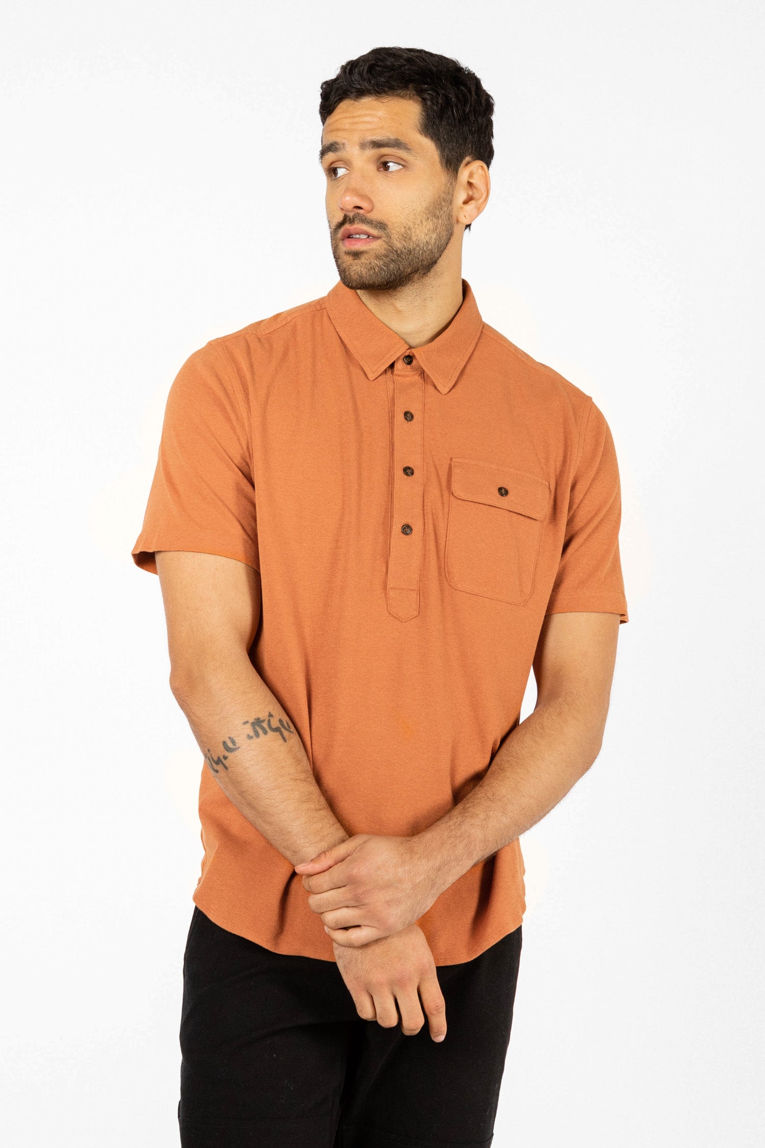 A man with short dark hair and a trimmed beard wears the Bridge & Burn Gary Knit Pullover in Clay Red Jersey with black pants. He stands against a plain white background, looking to his left with his hands clasped.