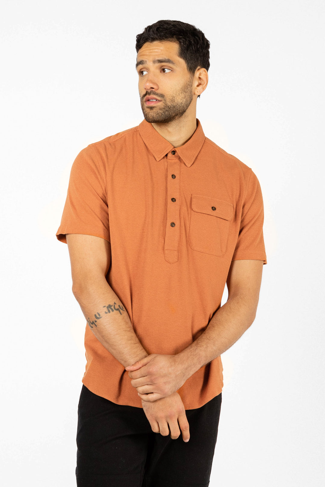 A man with short dark hair and a trimmed beard wears the Bridge & Burn Gary Knit Pullover in Clay Red Jersey with black pants. He stands against a plain white background, looking to his left with his hands clasped.