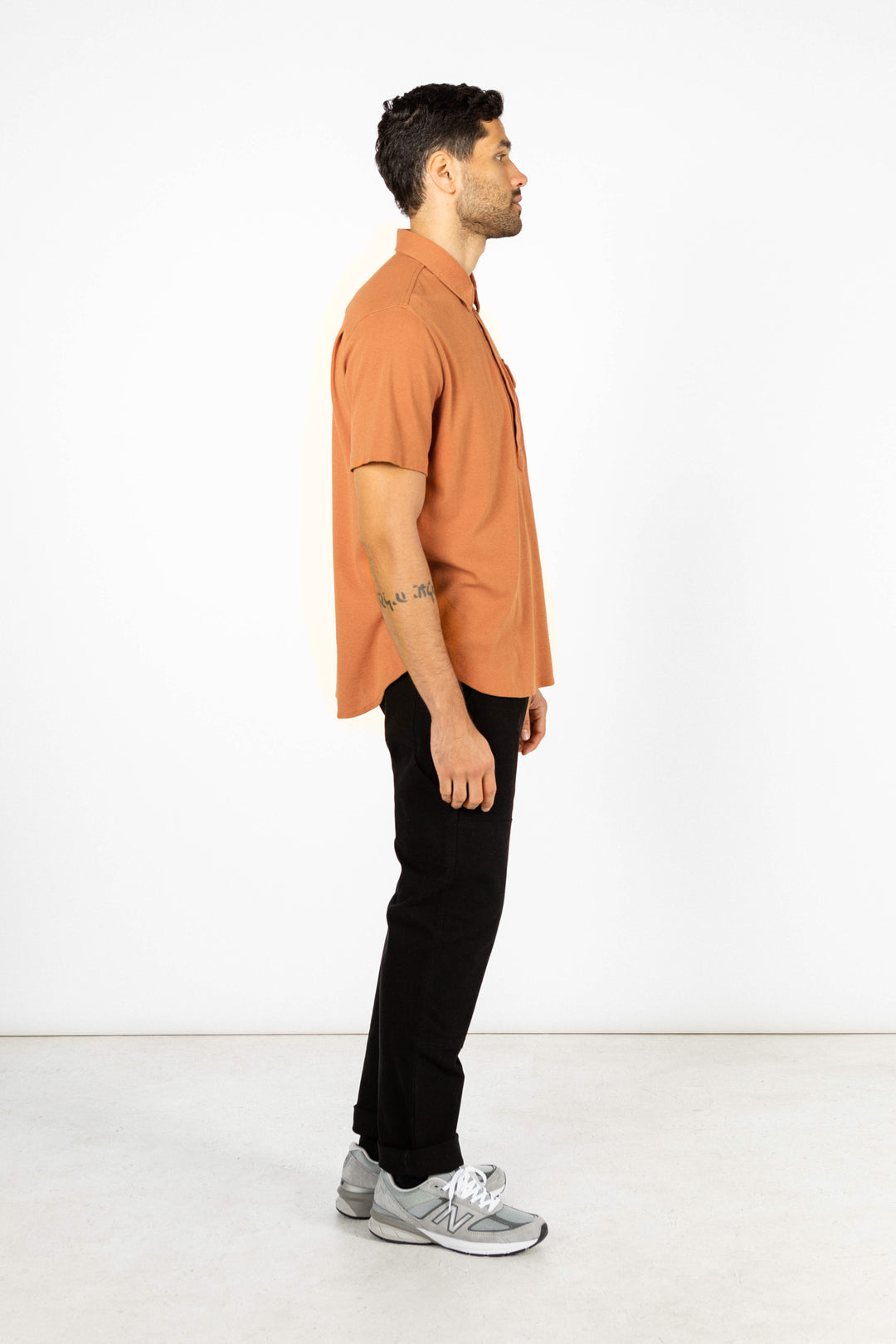 A man stands in profile against a white background, wearing the Bridge & Burn Gary Knit Pullover in Clay Red Jersey, paired with black pants and gray sneakers.