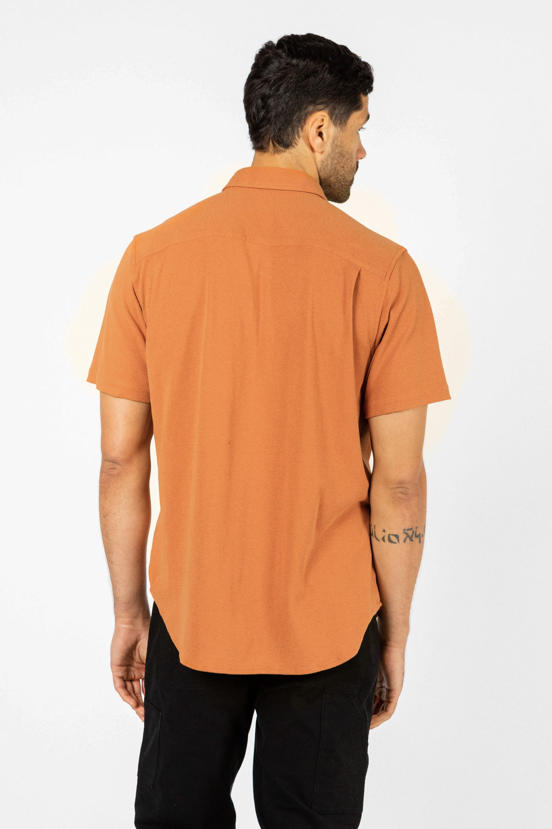 A man stands with his back to the camera, modeling Bridge & Burn’s Gary Knit Pullover in Clay Red Jersey and black pants against a plain white background.