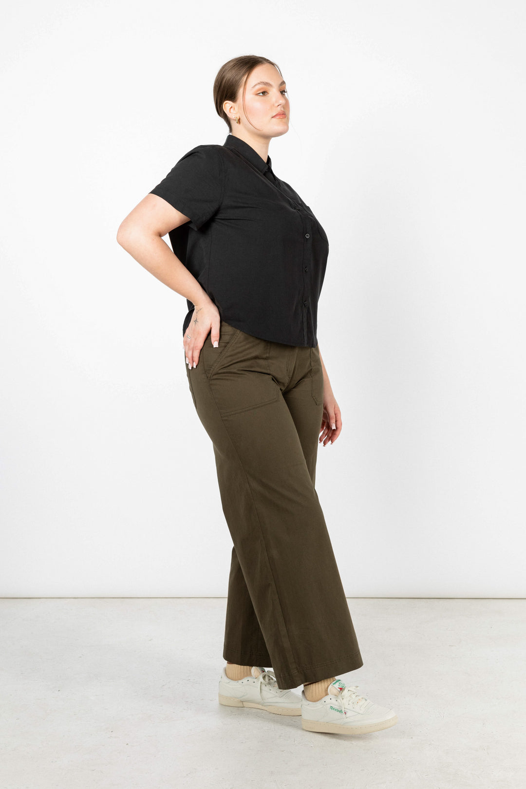 A woman poses sideways against a white backdrop in the Greer Cropped Shirt / Black Poplin by Bridge & Burn, styled with olive wide-leg pants and white sneakers. Her hair is tied back as she looks forward with confidence.
