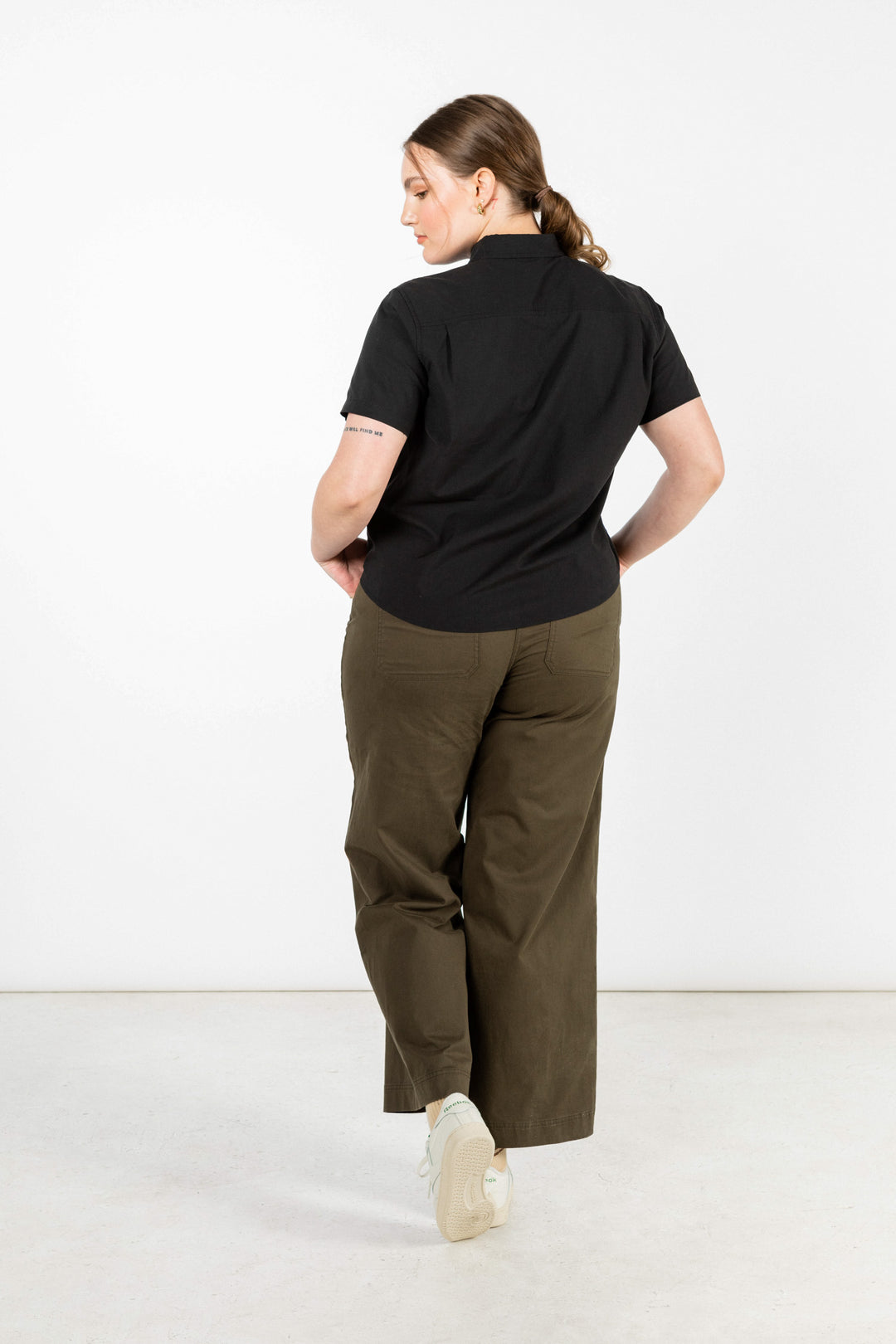 A person with light skin and brown hair in a low ponytail stands facing away, wearing the Bridge & Burn Greer Cropped Shirt in Black Poplin, olive green wide-leg pants, and white sneakers against a plain white background.
