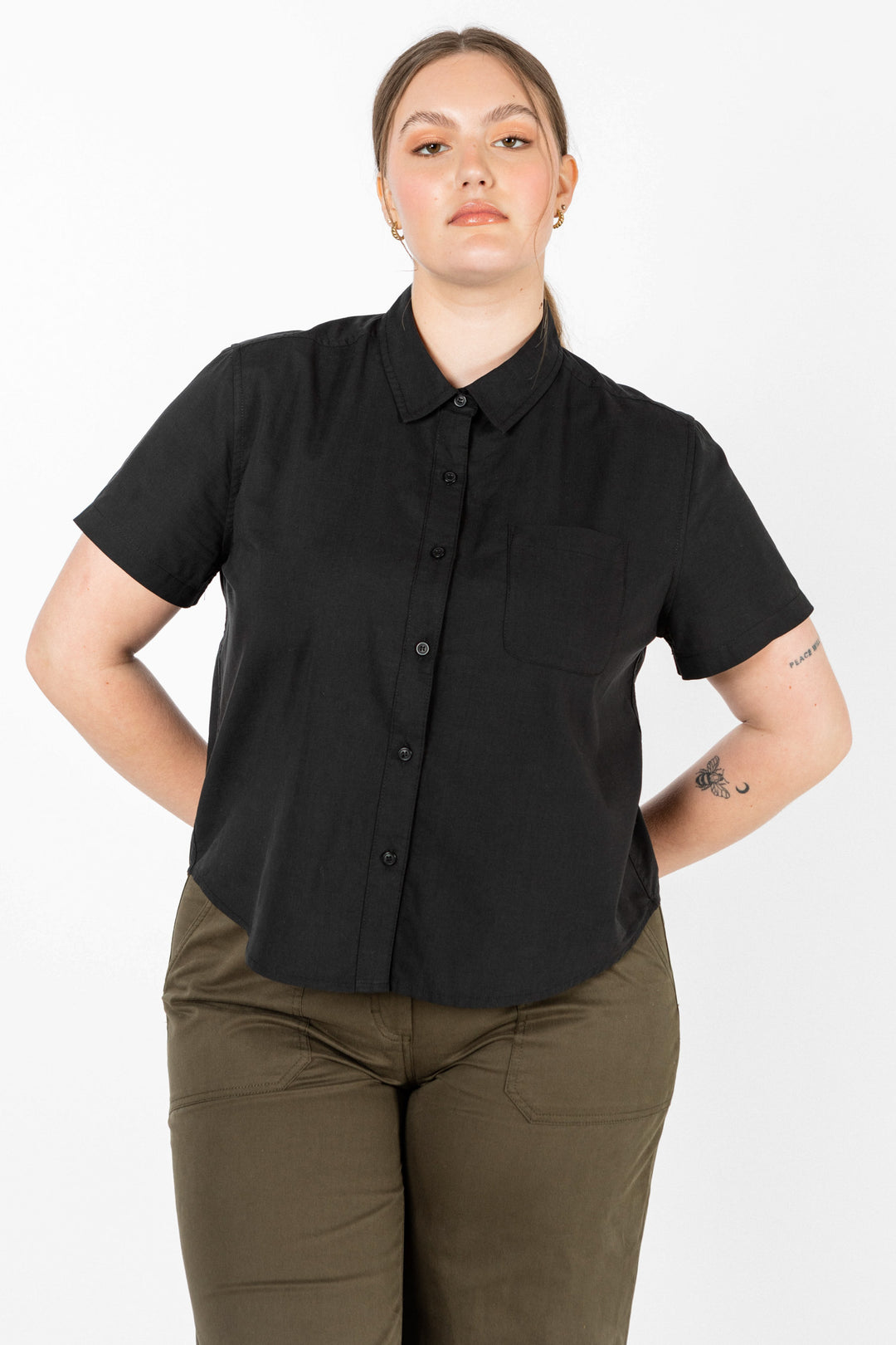 A person with light skin and long hair in a low ponytail wears the Bridge & Burn Greer Cropped Shirt in Black Poplin, paired with olive green pants, standing hands behind back against a plain white background.