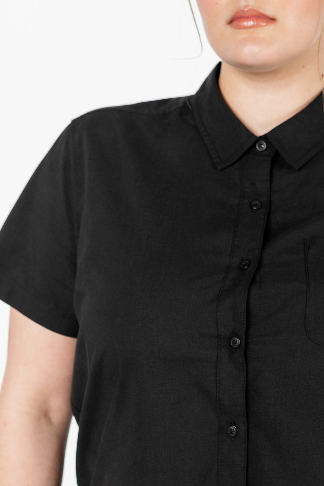 A person wears the Bridge & Burn Greer Cropped Shirt in black poplin—a short-sleeved, boxy fit button-up with a collar. The cropped image focuses on the upper torso and part of the face against a neutral background.