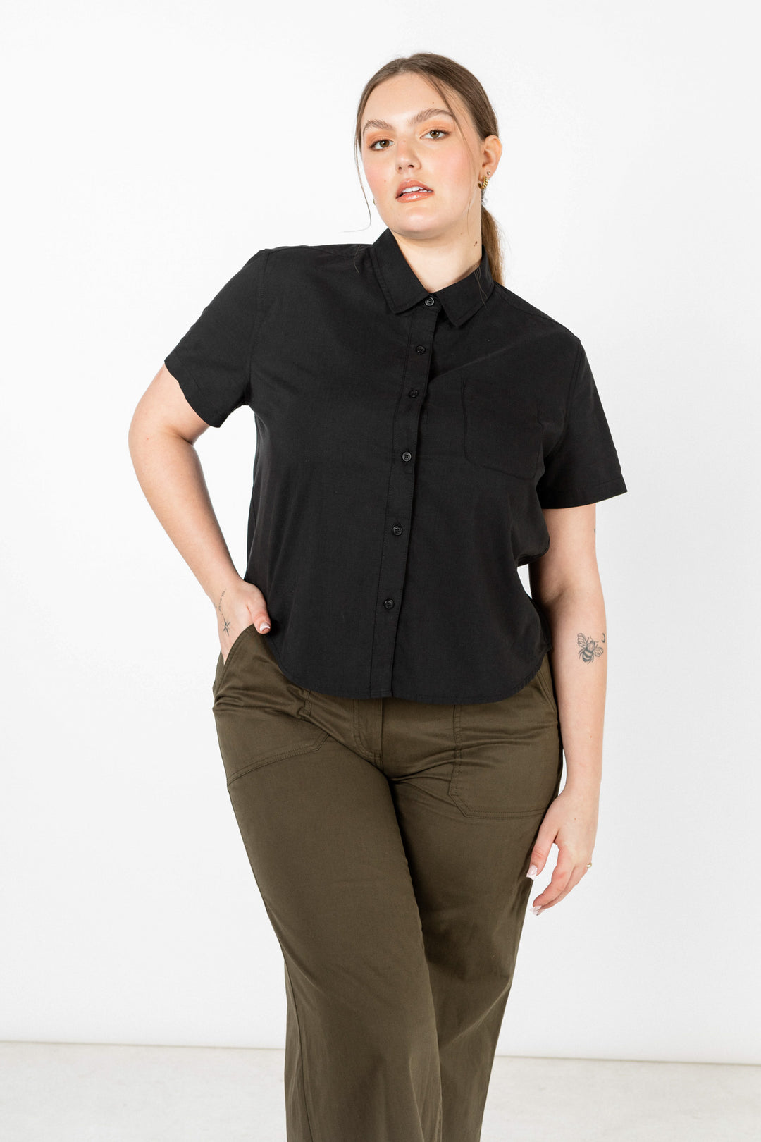 A woman with light brown hair pulled back stands against a white background, wearing the Bridge & Burn Greer Cropped Shirt in black poplin and olive green pants, showing a small butterfly tattoo on her left forearm.