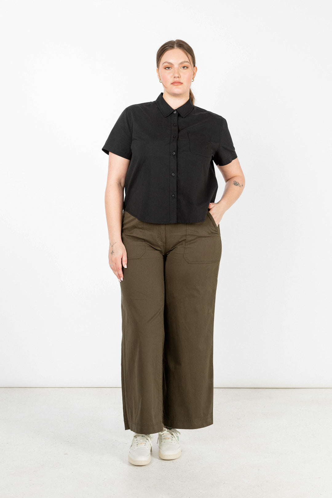 Against a white background, a person with light brown hair pulled back wears Bridge & Burn's Greer Cropped Shirt in black poplin, olive green wide-leg pants, and white sneakers—one hand is in their pocket.