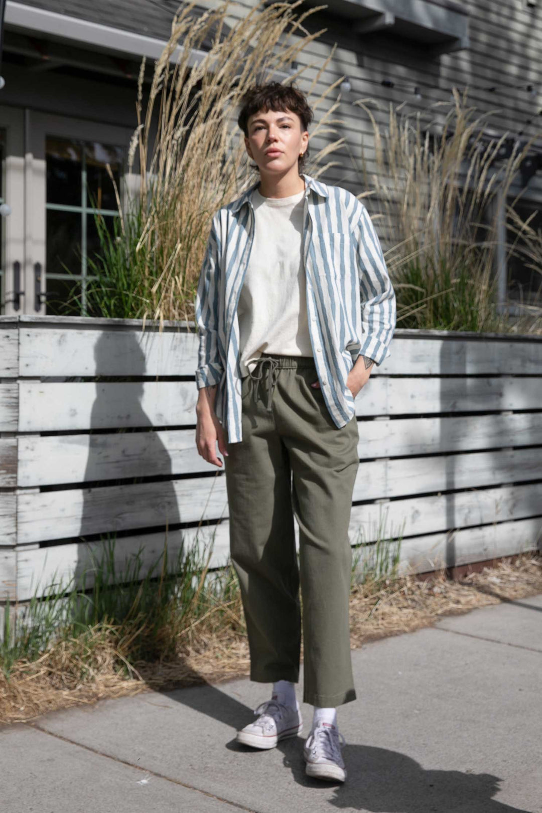 A person with short hair stands outside by tall grasses and a wooden fence, wearing the Bridge & Burn Haley Shirt in Beach Stripe over a white T-shirt, paired with green pants and white sneakers.