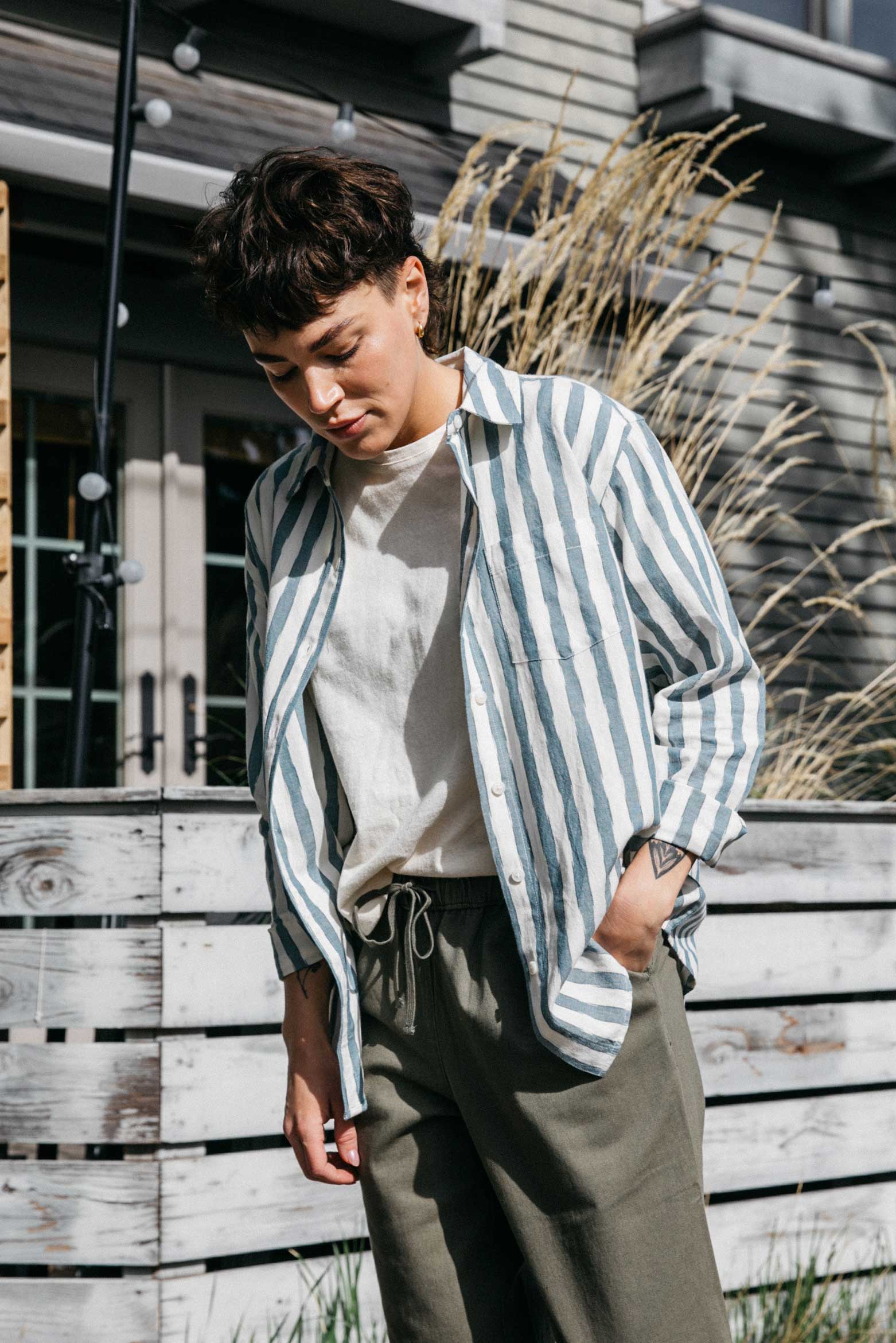 Wearing the Bridge & Burn Haley Shirt in Beach Stripe, a person stands outdoors by a wooden fence, looking down with one hand in their pocket. Tall grass and a building are visible in the background.
