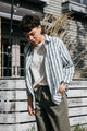 Wearing the Bridge & Burn Haley Shirt in Beach Stripe, a person stands outdoors by a wooden fence, looking down with one hand in their pocket. Tall grass and a building are visible in the background.