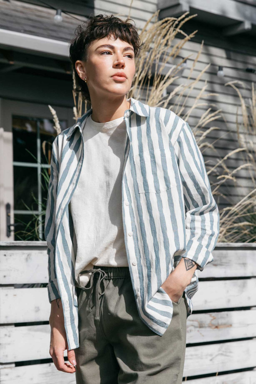 Standing outdoors by tall grass and a wooden fence, a person in the Bridge & Burn Haley Shirt in Beach Stripe over a white tee and green pants soaks up the sun, one hand in their pocket, their short hair catching the light.