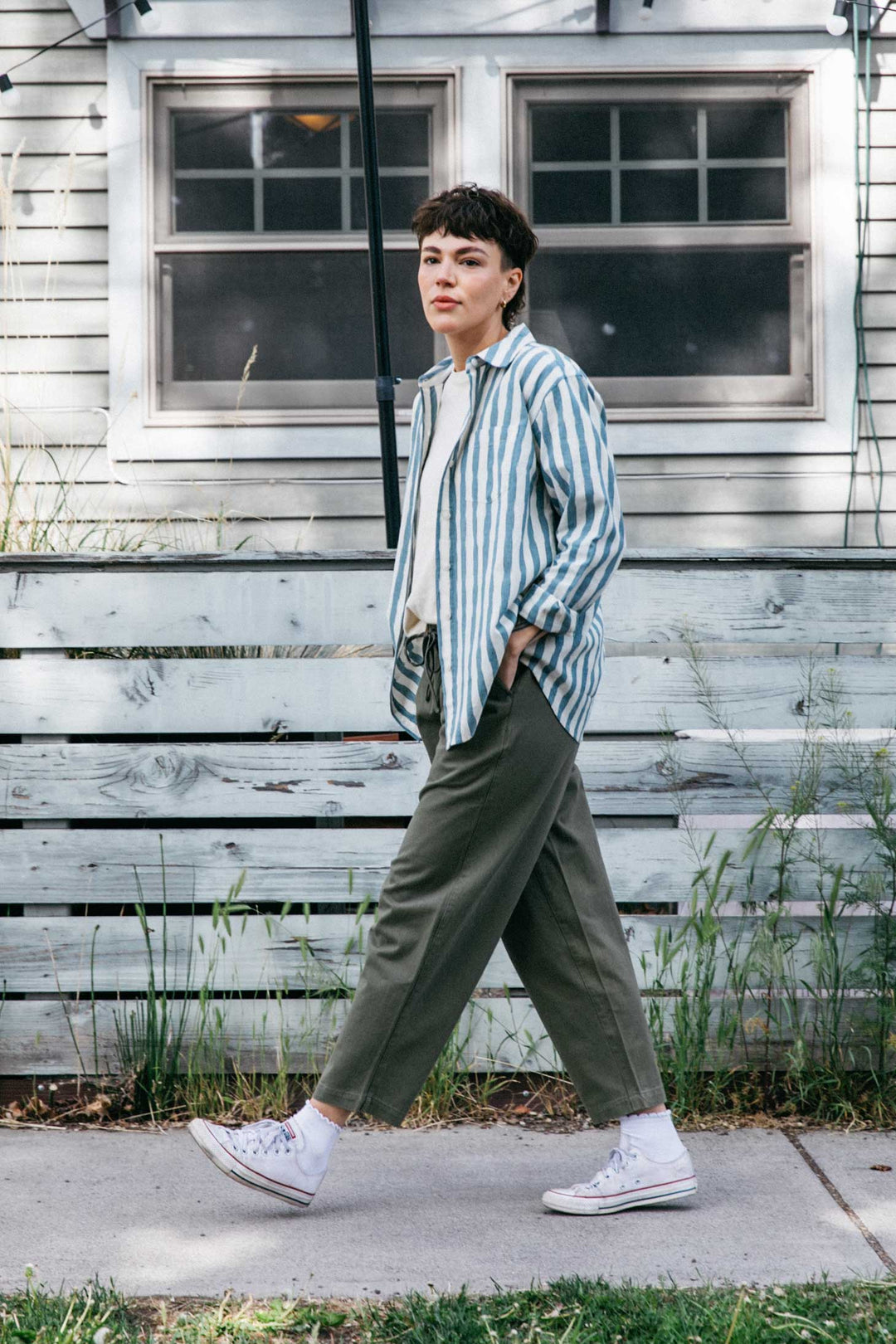 A person with short dark hair walks outside by a light wooden fence, wearing the Bridge & Burn Haley Shirt in Beach Stripe, paired with olive green pants, white socks, and white sneakers.