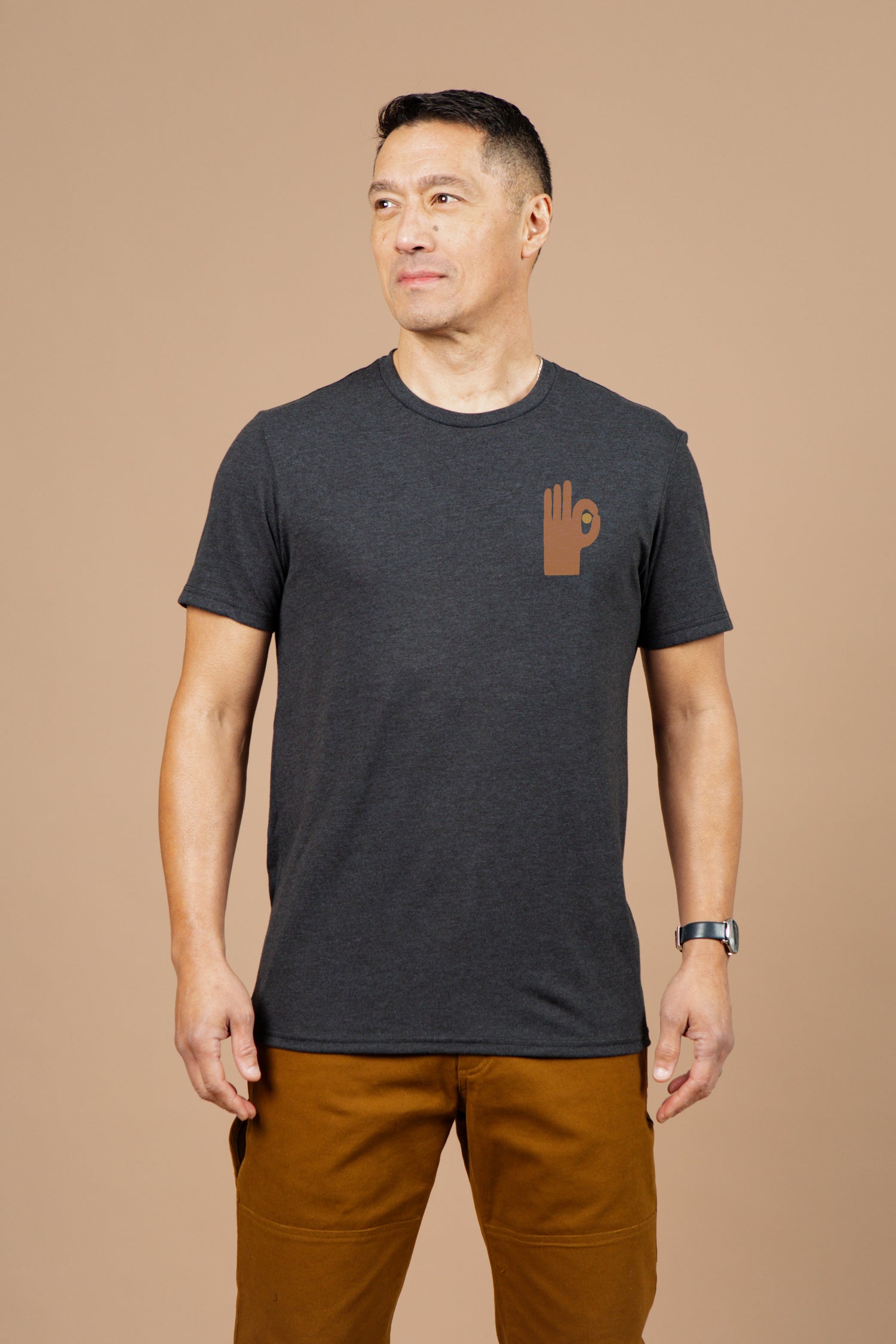 A man stands against a beige background wearing the Bridge & Burn Men's Hand Eye Tee in black, featuring a brown hand symbol on the left chest. He pairs it with brown pants.