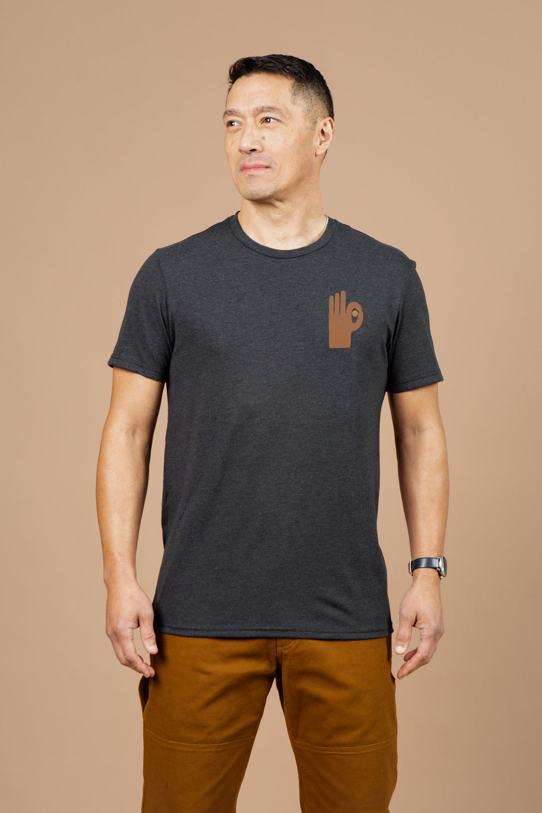 A man stands against a beige background wearing the Bridge & Burn Men's Hand Eye Tee in black, featuring a brown hand symbol on the left chest. He pairs it with brown pants.