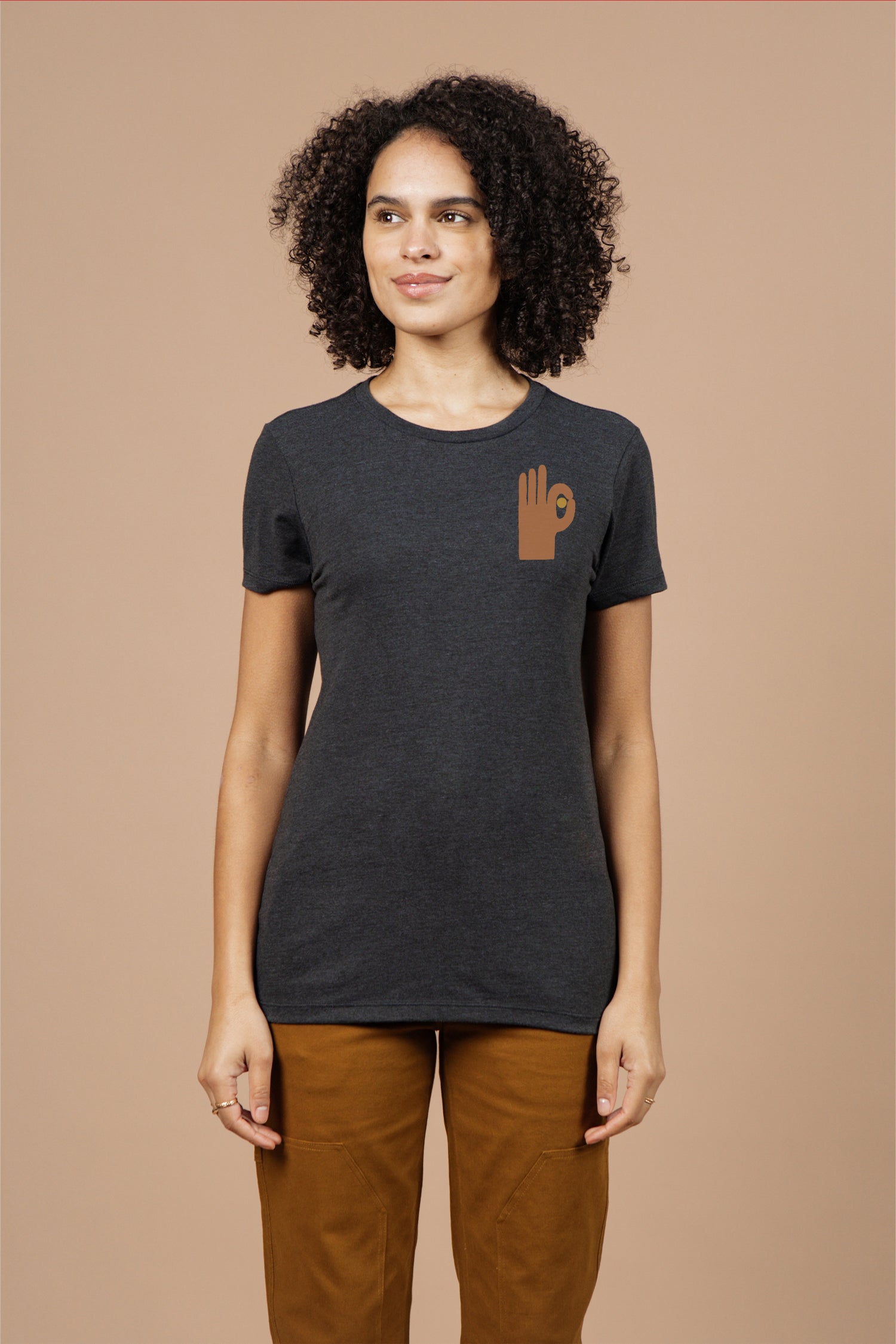 A woman with curly hair wears the Bridge & Burn Women's Hand Eye Tee in black, featuring a brown hand making an OK gesture on the left chest, paired with brown pants, standing against a beige background.