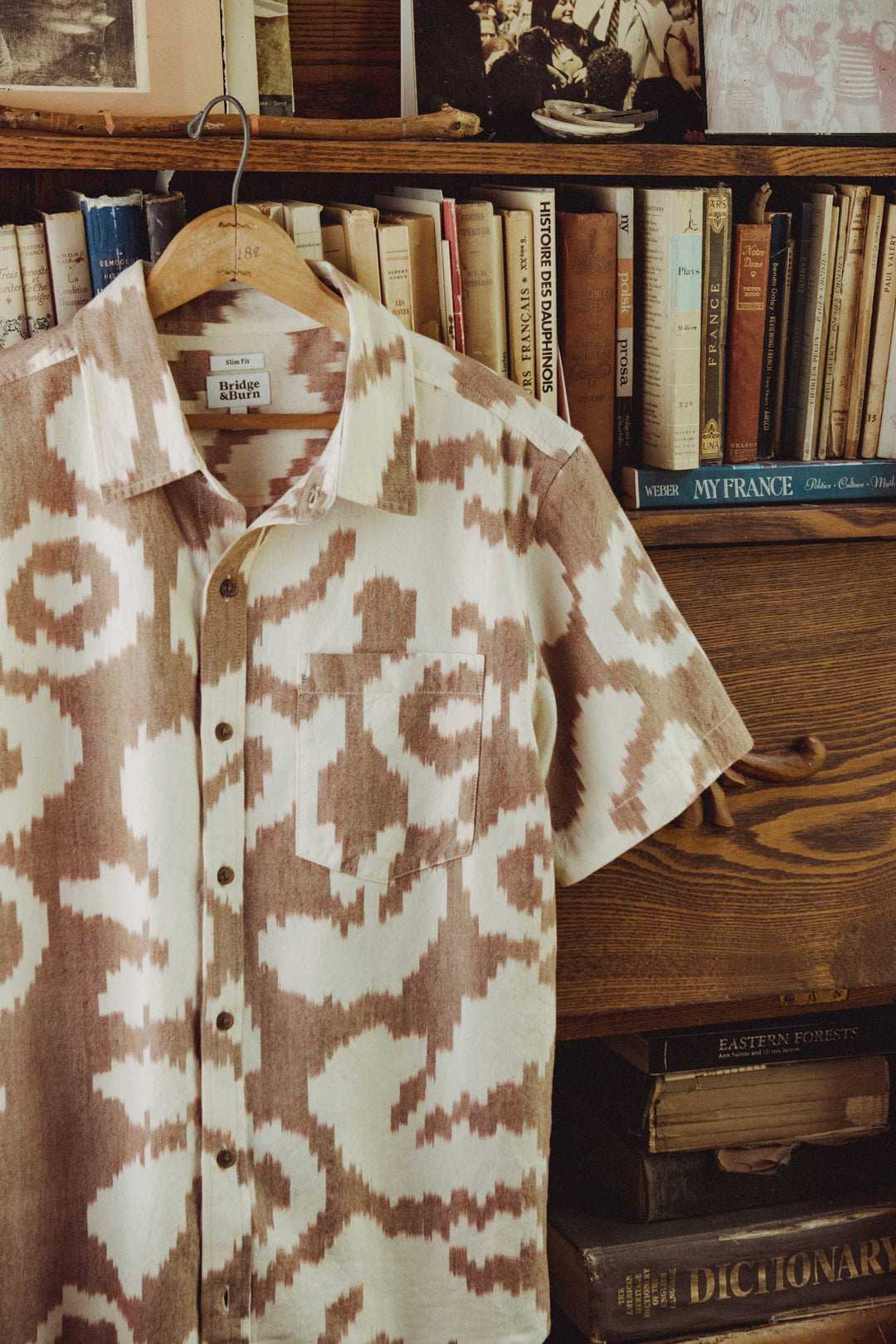 The Bridge & Burn Harbor Slim Shirt / Pop Leaf, a beige and brown summer-weight cotton shirt with a patterned design, hangs on a hanger in front of a wooden bookshelf filled with books and vintage photos, with stacked books visible below.