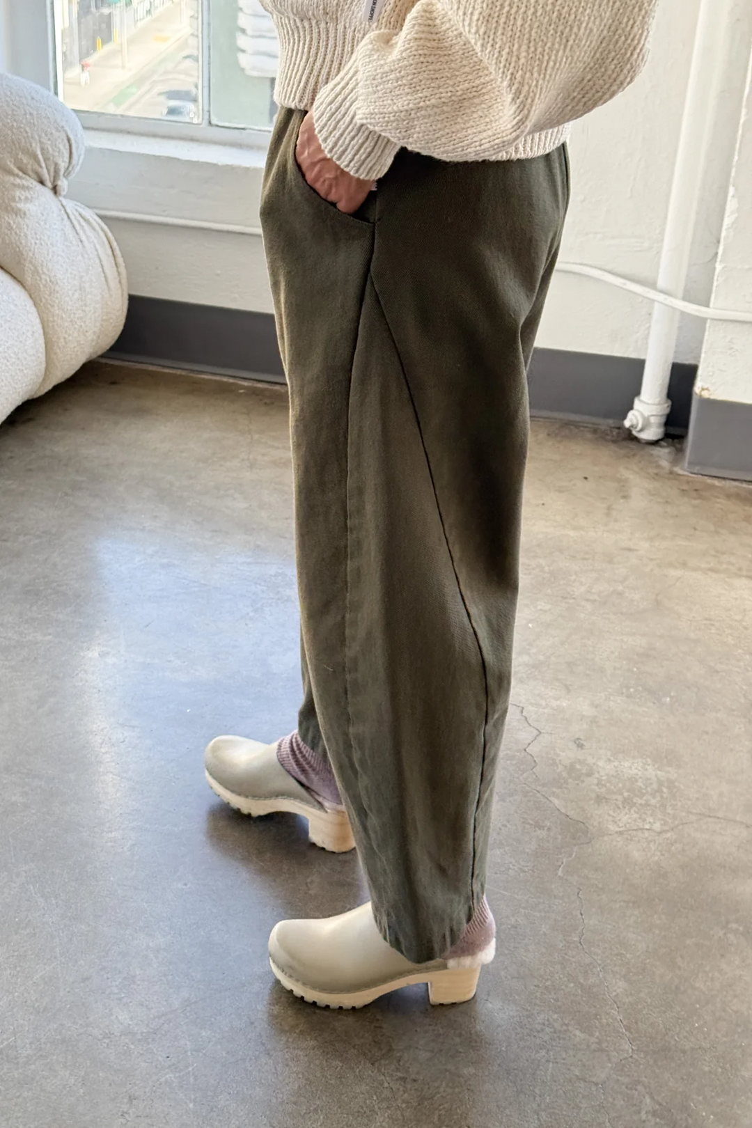 A person stands indoors on concrete, wearing Le Bon Shoppe Arc Pant in Olive with a cream knit sweater, light socks, and off-white clogs. Hand in pocket; natural light streams through a nearby window.