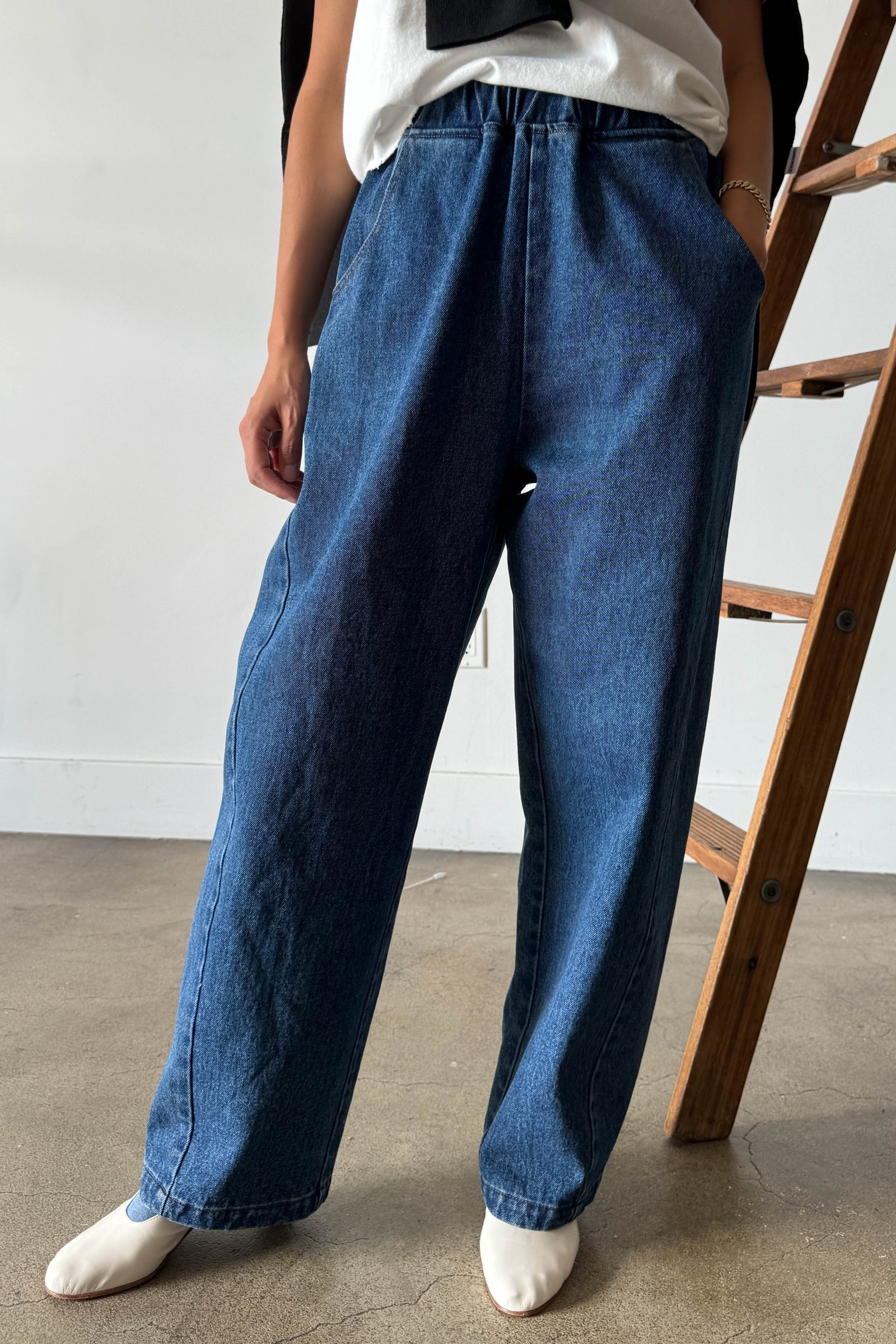 A person stands indoors on a concrete floor near a wooden ladder, wearing the Le Bon Shoppe - Long Arc Pant / Blue Denim by Le Bon Shoppe—loose-fitting blue jeans crafted from heavyweight denim—paired with a white shirt and white pointed shoes. The upper part of their face is not visible.