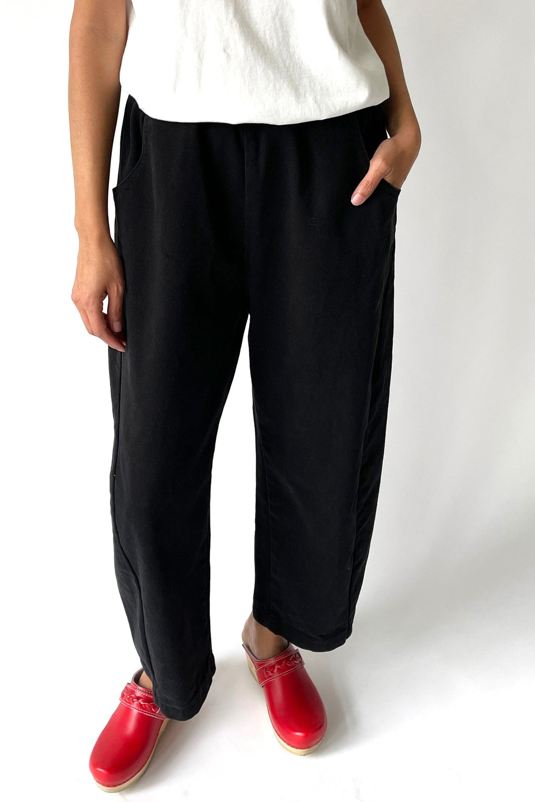 A person wearing the Le Bon Shoppe - Arc Pant in Black by Le Bon Shoppe stands against a white background with one hand in their pocket. Only the lower half of their body is visible, styled with a white top and red clogs.