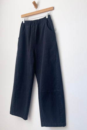 A pair of Le Bon Shoppe - Arc Pant / Black from Le Bon Shoppe, featuring a wide-leg silhouette in heavyweight canvas with an elastic waistband and side pockets, hangs on a wooden hanger against a white wall.