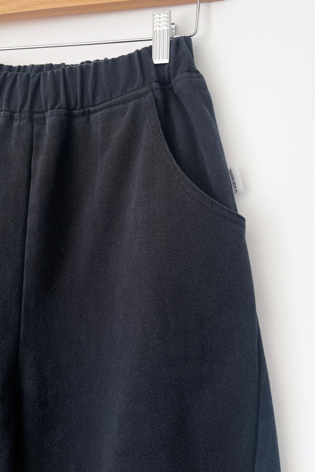 Close-up of the Le Bon Shoppe - Arc Pant in black, crafted from heavyweight canvas, hanging on a wooden hanger. The image highlights the elastic waistband, front pocket, and a small fabric tag on the side, all set against a white background.