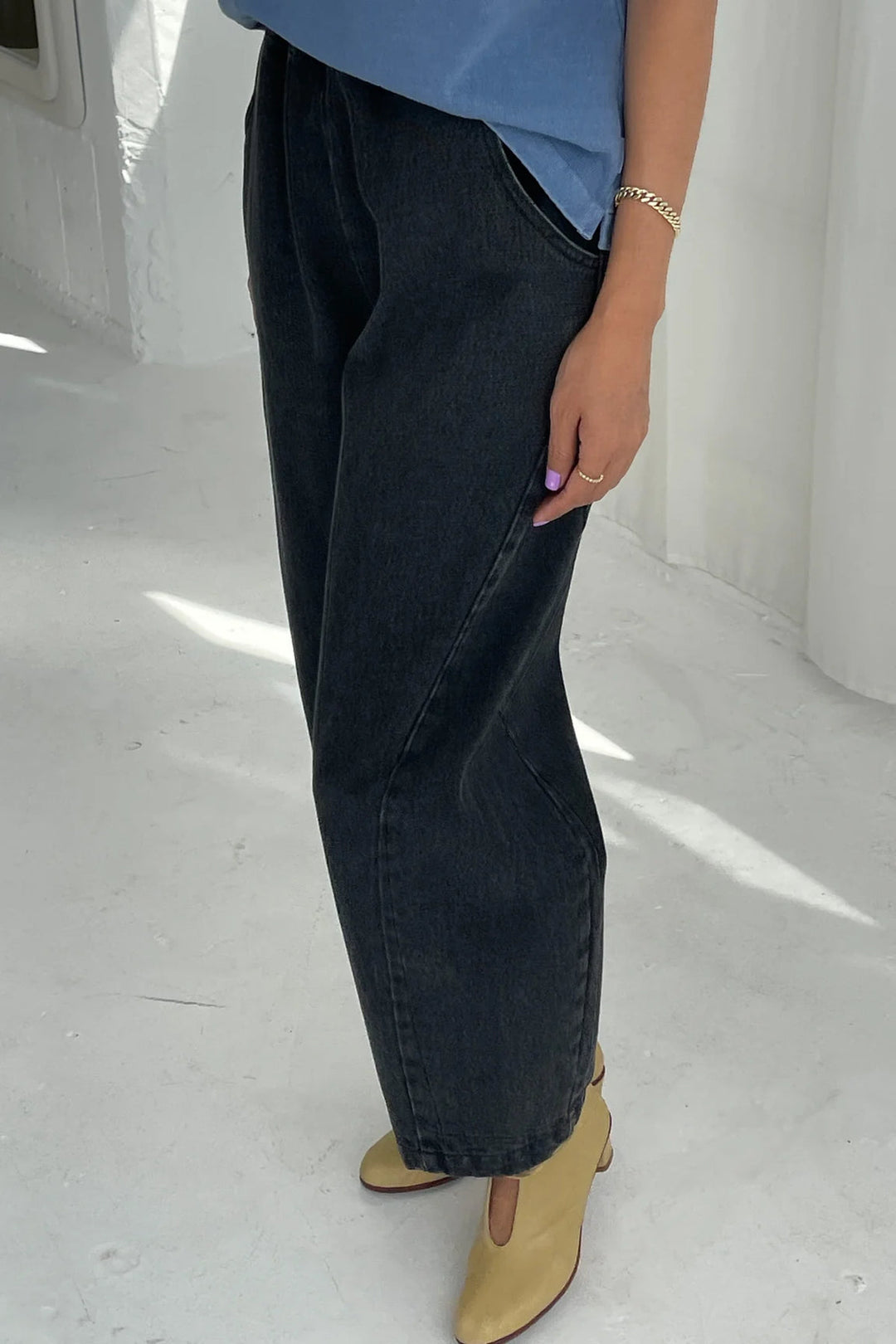 A person stands indoors wearing Le Bon Shoppe’s Arc Pant in Black Denim, paired with a blue top and beige heeled shoes. Their nails are painted lavender, and a gold bracelet adorns their wrist. Only the lower half of their body and one arm are visible.