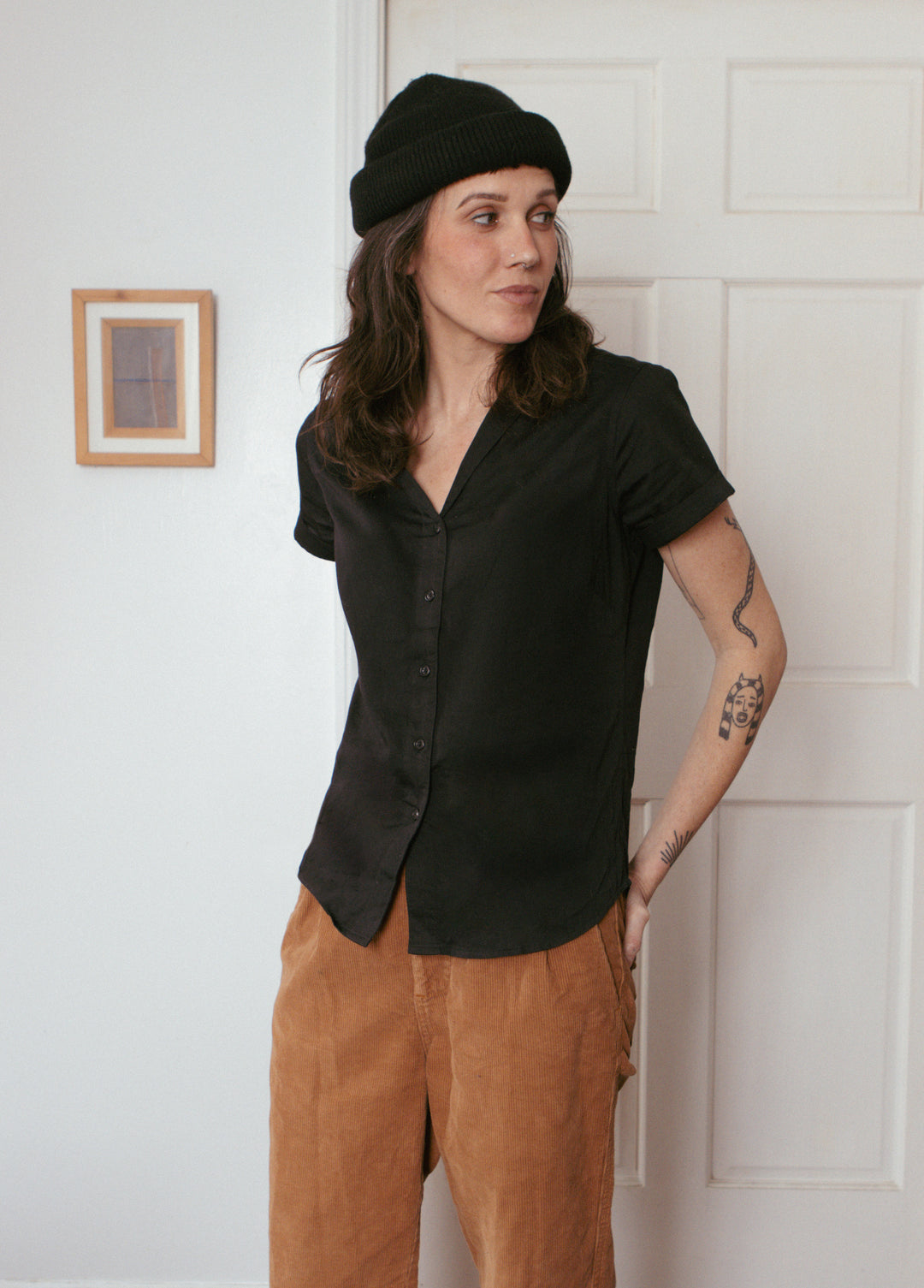 A person with long brown hair and a black beanie wears the Bridge & Burn Innes Shirt in black—short sleeves, relaxed fit—paired with brown pants, standing indoors by a white door, hand in pocket and tattoo visible on their right arm.