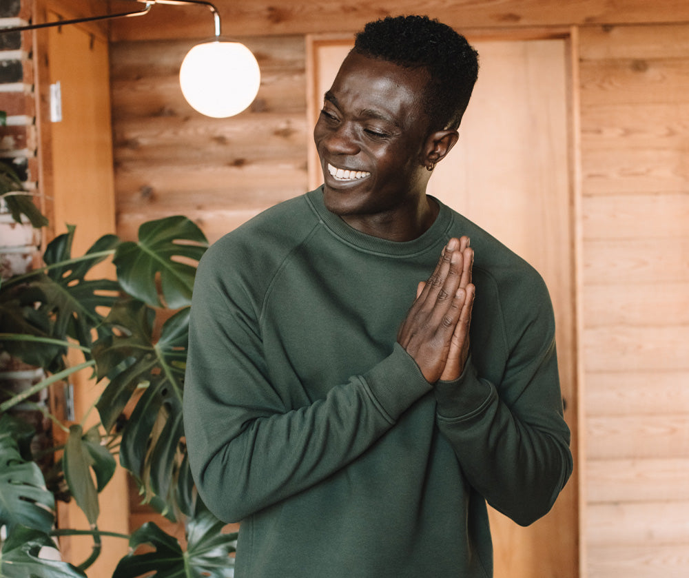 A smiling man in a green sweatshirt stands indoors, hands clasped together, with a large green plant and wooden walls in the background.