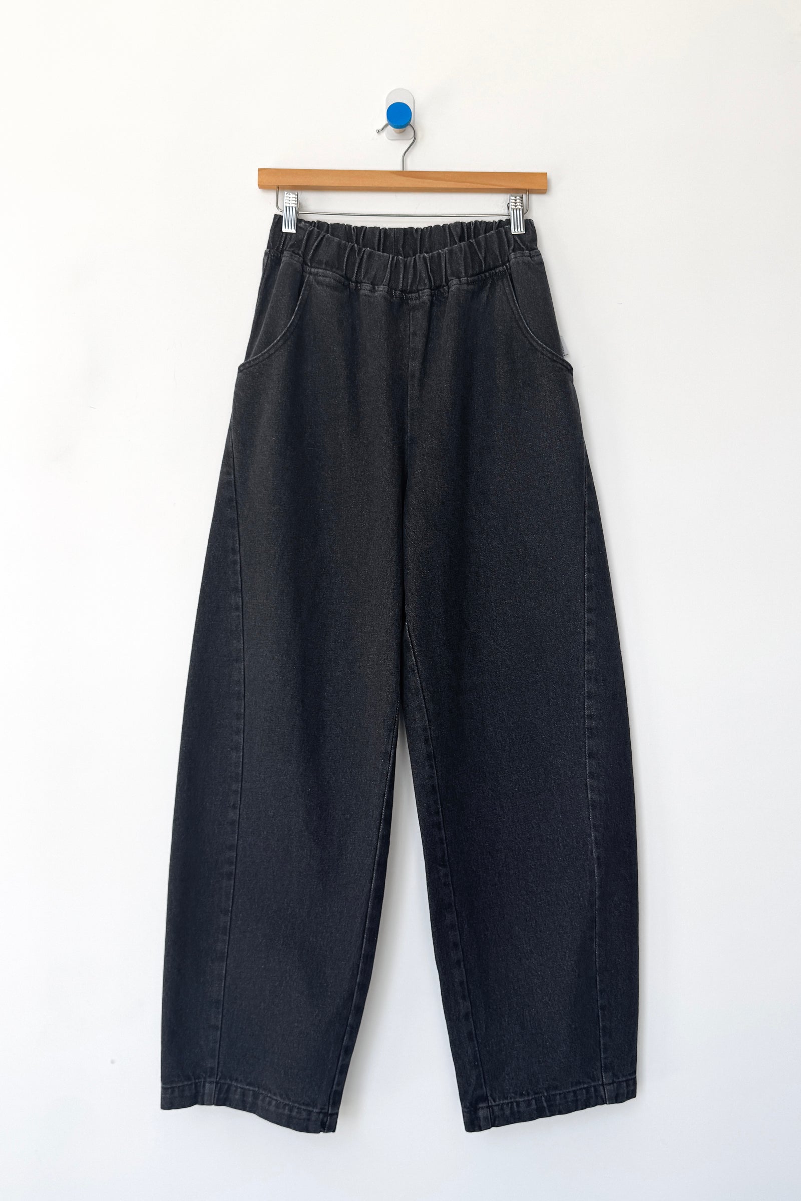 A pair of Le Bon Shoppe - Long Arc Pant in black denim, featuring a loose fit, elastic waistband, and crafted from heavyweight denim, hangs on a wooden hanger against a white wall.