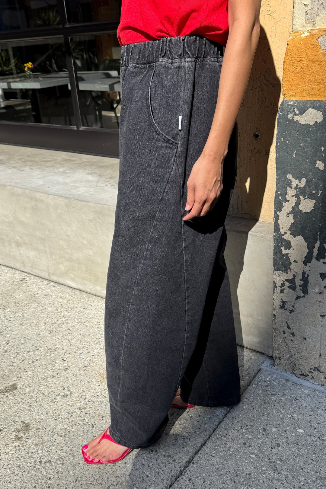 A person is wearing Le Bon Shoppe’s Long Arc Pant in Black Denim, featuring visible seams and an extended inseam. Only the lower body is visible as they stand outdoors on a sunny day beside a yellow and black textured wall.