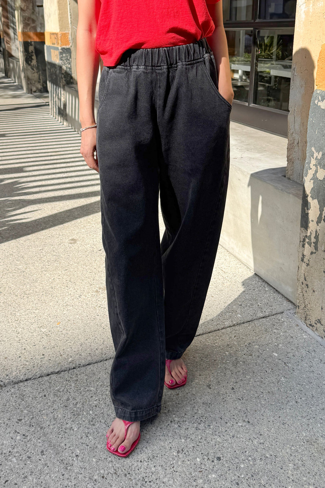A person is seen standing on a sunny sidewalk with one hand in their pocket, wearing Le Bon Shoppe’s Long Arc Pant in black denim. Only their lower body is visible.