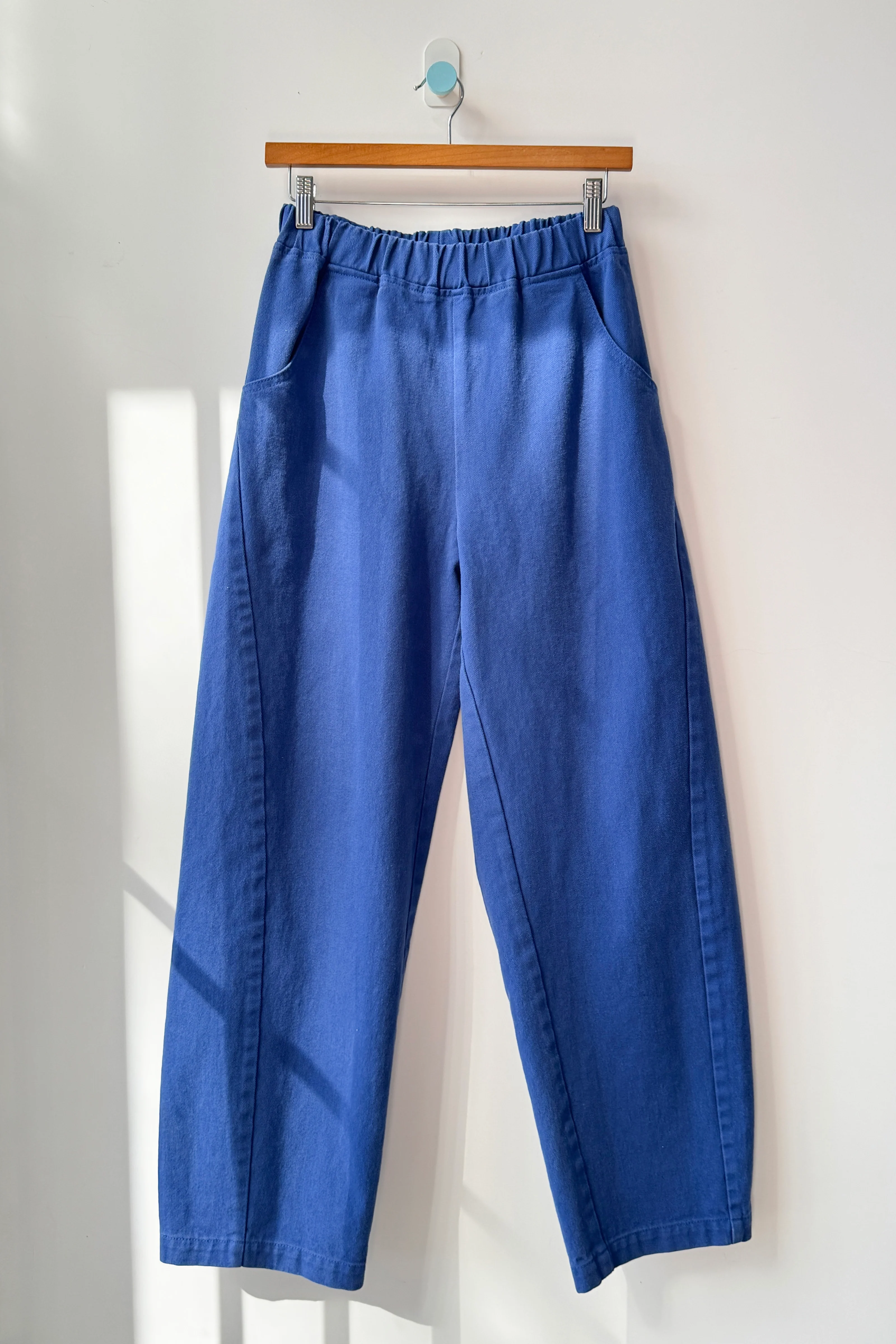 The Le Bon Shoppe Long Arc Pant in Azure, crafted from heavy-weight denim with a loose fit, high waist, and elastic waistband, hangs on a wooden hanger against a white wall as sunlight casts soft shadows.