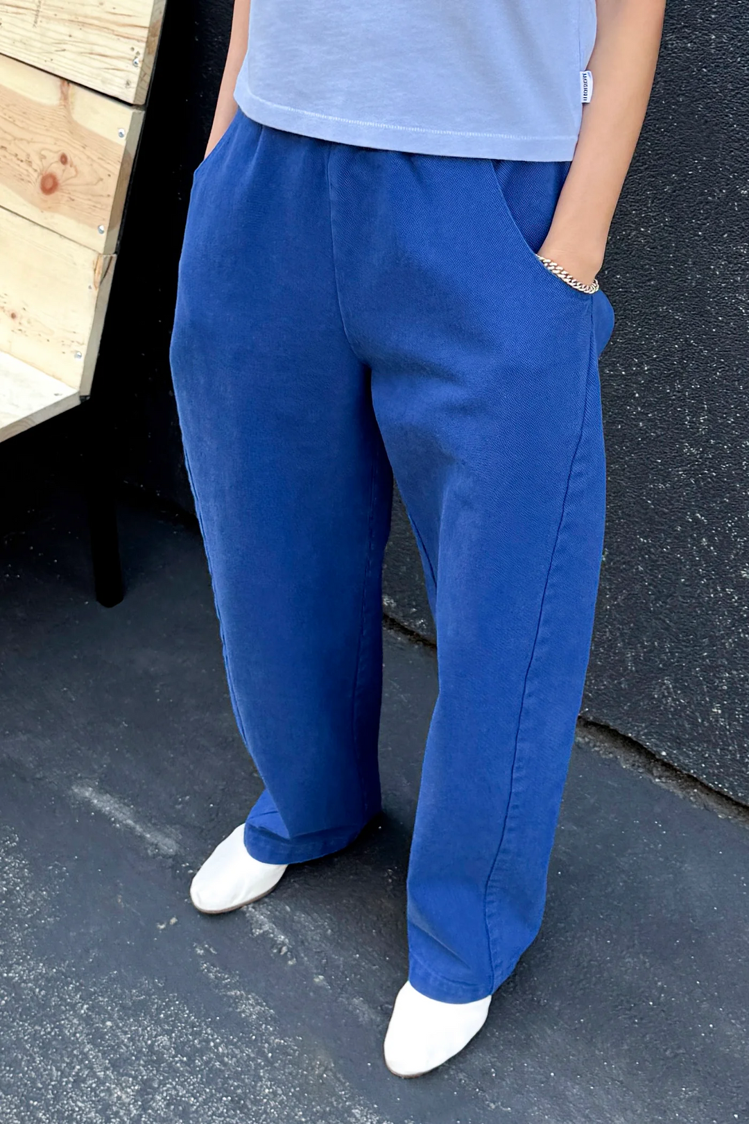 Someone wearing Le Bon Shoppe's Long Arc Pant in Azure, paired with a light grey top and white shoes, stands on a blacktop surface with hands in pockets. A wooden structure appears on the left.