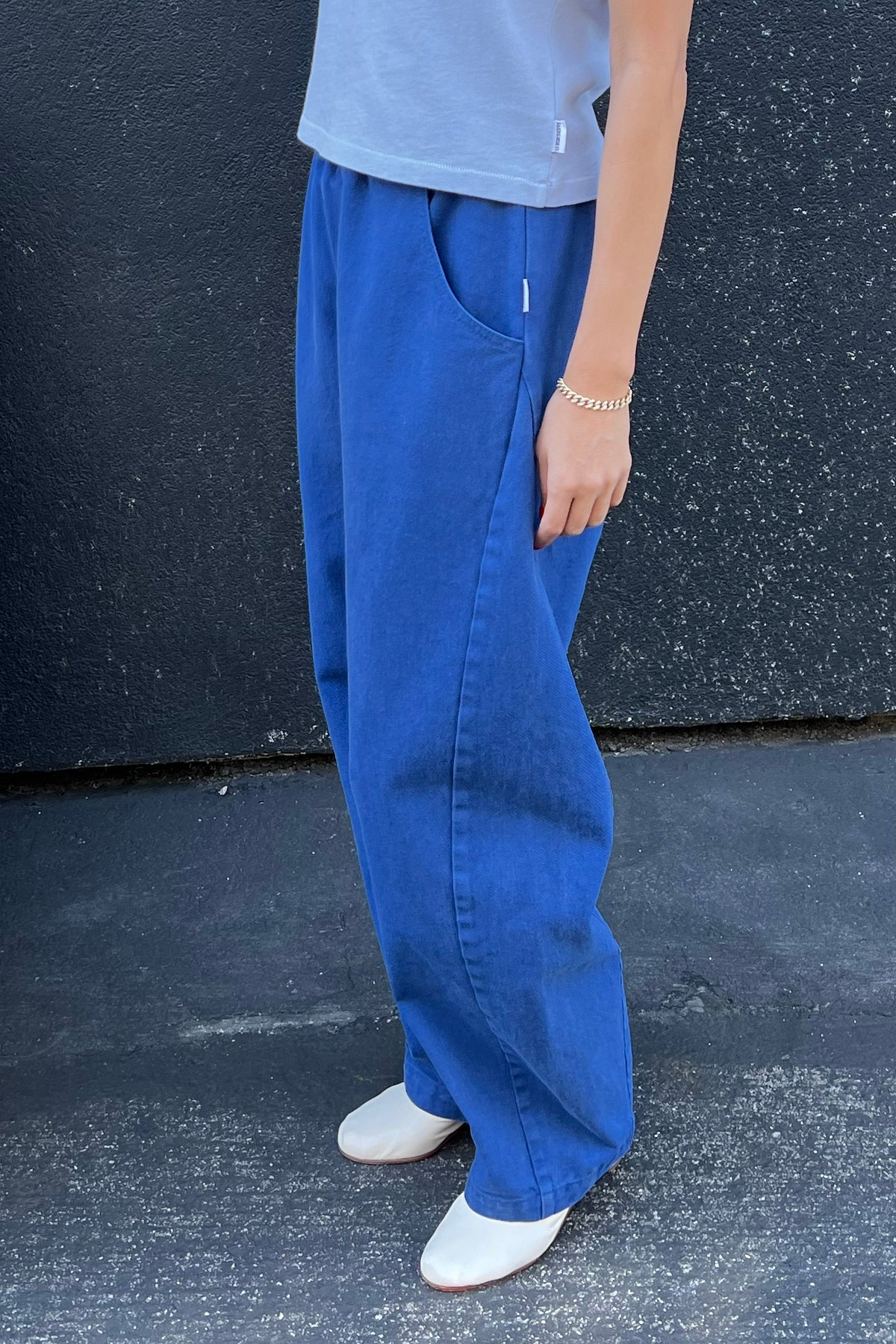 A person wears Le Bon Shoppe's Long Arc Pant in Azure, paired with a light gray top, white shoes, and a gold bracelet, standing against a dark textured wall and pavement.
