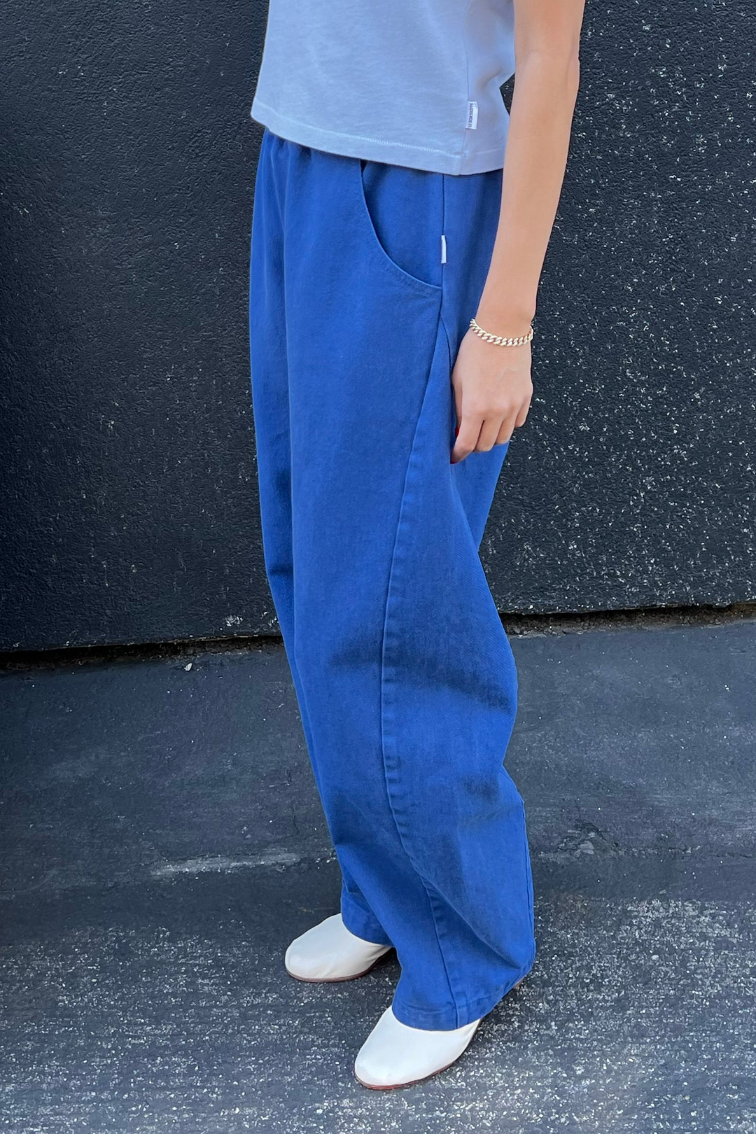 A person wears Le Bon Shoppe's Long Arc Pant in Azure, paired with a light gray top, white shoes, and a gold bracelet, standing against a dark textured wall and pavement.