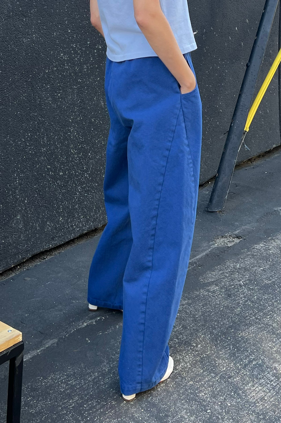 Wearing Le Bon Shoppe's Long Arc Pant in Azure, a person with hands in pockets stands on a paved surface beside a dark textured wall and metal pole, pairing the pants with a light blue top.