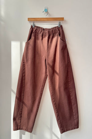The Le Bon Shoppe - Long Arc Pant in Chocolate, made from heavyweight denim with a high waist, elastic waistband, and side pockets, hangs on a wooden hanger against a sunlit white wall.