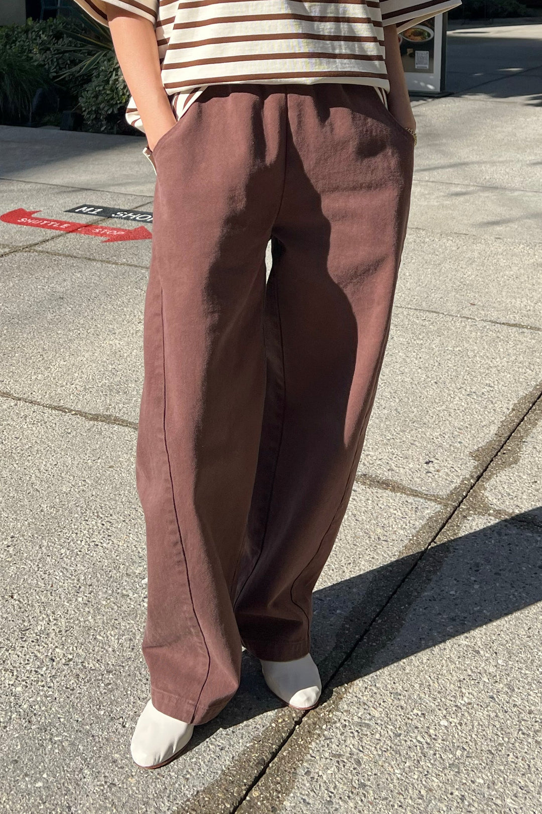 A person stands outdoors in wide-legged Le Bon Shoppe - Long Arc Pant / Chocolate, paired with a striped short-sleeve shirt and white shoes. With hands in pockets and face unseen, sunlight casts shadows on the pavement.