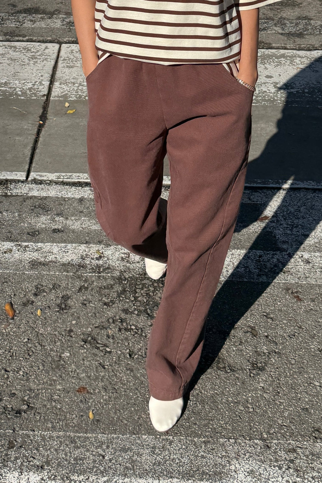 Person stands on a street crosswalk wearing Le Bon Shoppe - Long Arc Pant / Chocolate with an extended inseam, a striped shirt, and white pointed shoes, hands in pockets. Their upper face is not visible.