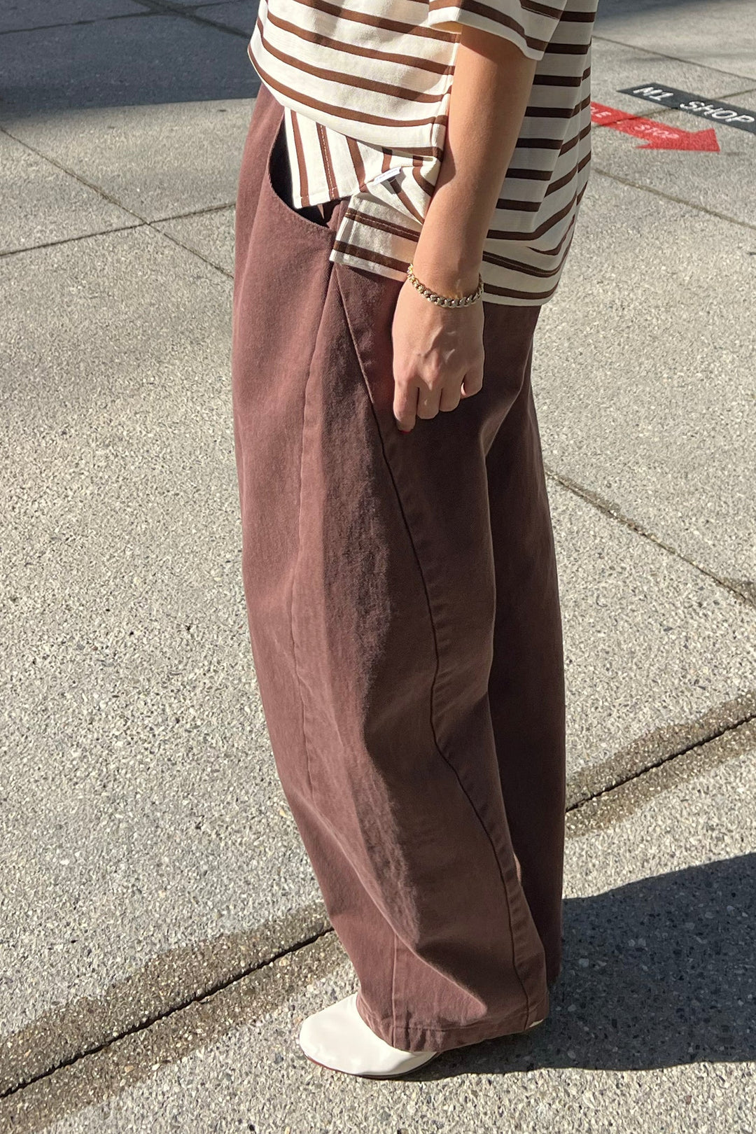 A person stands on a sunlit sidewalk wearing Le Bon Shoppe - Long Arc Pant / Chocolate, a striped beige and brown top, white shoes, and a chain bracelet. Their face is not visible.