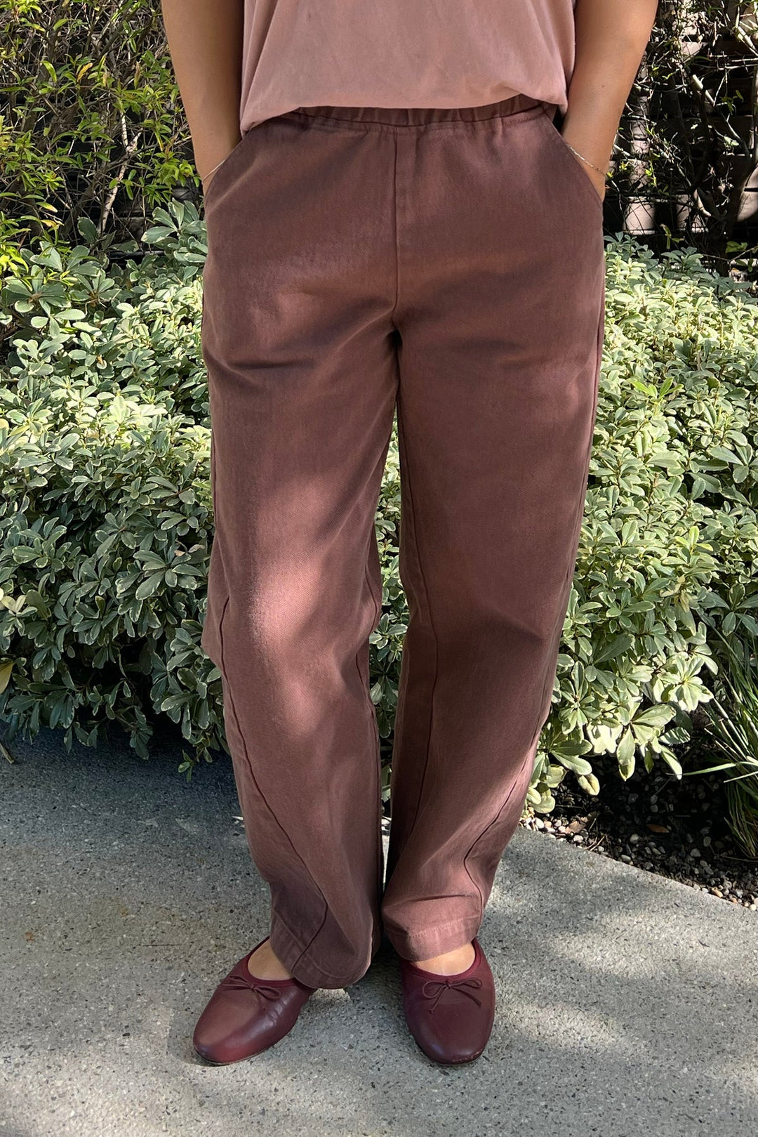 Someone stands outdoors with hands in pockets, wearing Le Bon Shoppe - Long Arc Pant in Chocolate and matching brown flat shoes. Green leafy plants are in the background, and their upper body is partially visible.