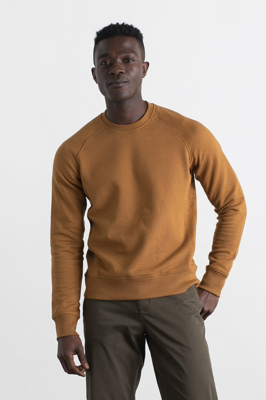 A man stands against a white background, wearing the Bridge & Burn Fremont Crew Sweatshirt in Bronze and olive green pants. He faces the camera with a neutral expression and has one hand in his pocket.