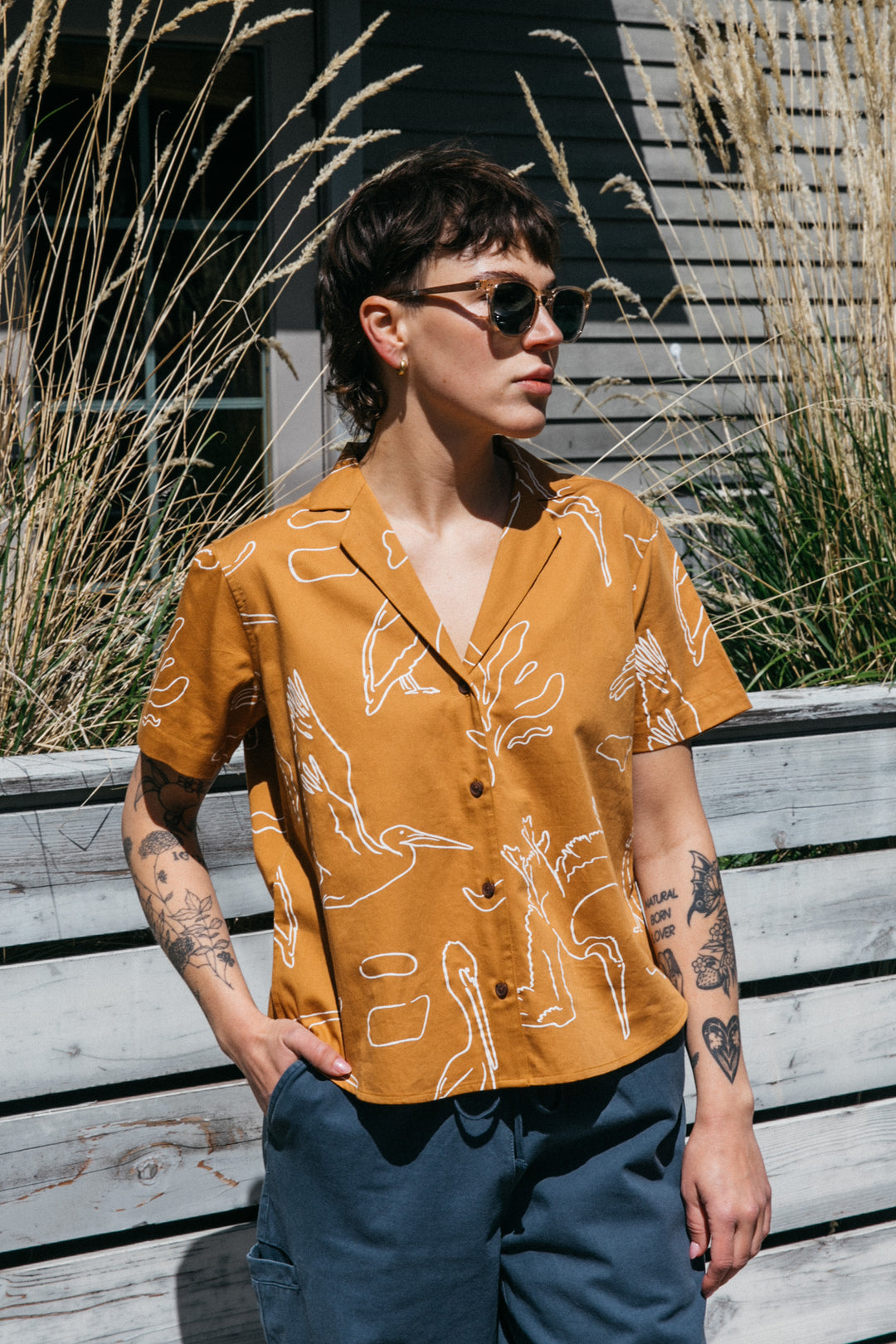 A person with short dark hair, sunglasses, and tattoos wears the Bridge & Burn Mila Cropped Shirt in Shoreflight and blue pants. They stand outside under sunlight, in front of tall grass and a wooden fence.