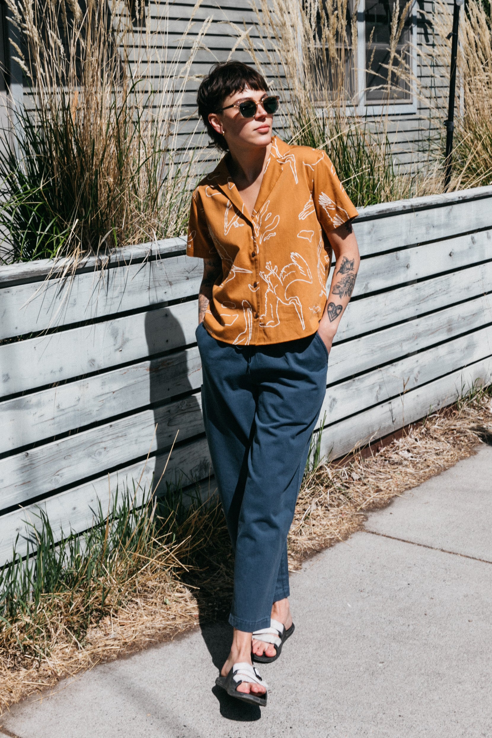 A person in the Bridge & Burn Mila Cropped Shirt / Shoreflight, sunglasses, blue pants, and sandals stands by a wooden fence and tall grass on a sidewalk, leaning back with hands in pockets.