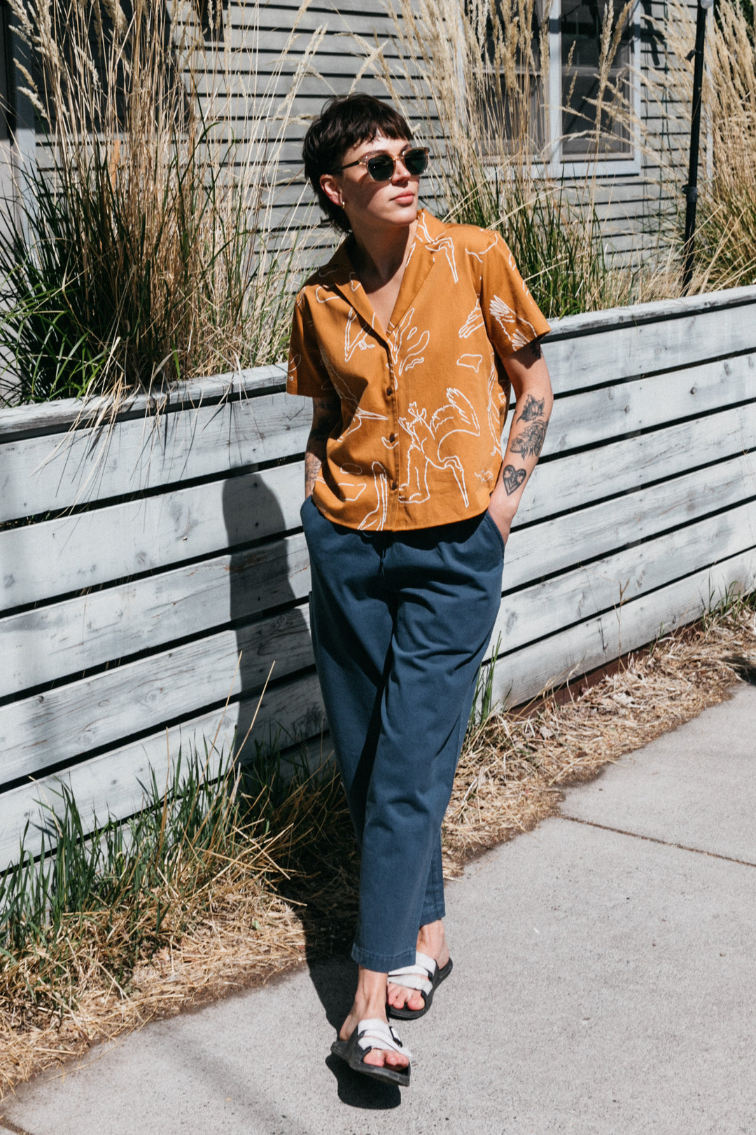 A person in the Bridge & Burn Mila Cropped Shirt / Shoreflight, sunglasses, blue pants, and sandals stands by a wooden fence and tall grass on a sidewalk, leaning back with hands in pockets.