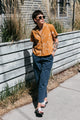 A person in the Bridge & Burn Mila Cropped Shirt / Shoreflight, sunglasses, blue pants, and sandals stands by a wooden fence and tall grass on a sidewalk, leaning back with hands in pockets.