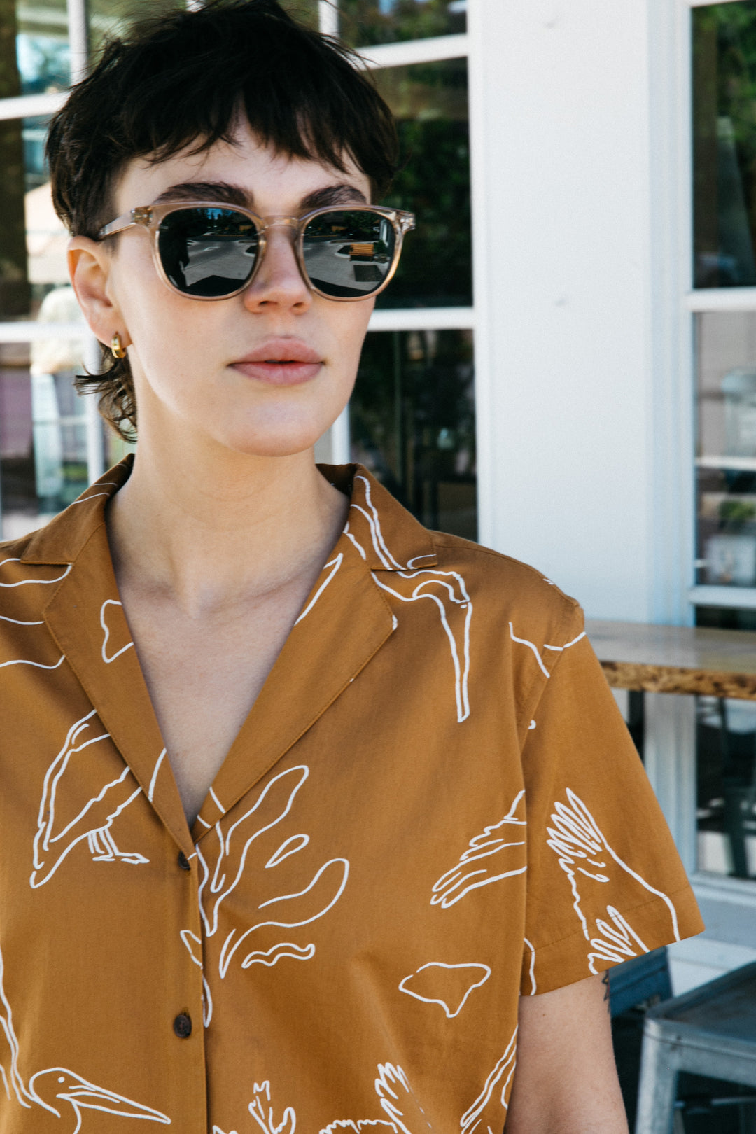 A person with short dark hair and round sunglasses wears the Bridge & Burn Mila Cropped Shirt in Shoreflight, a relaxed-fit brown button-up with white bird designs, while standing outdoors near a windowed building.